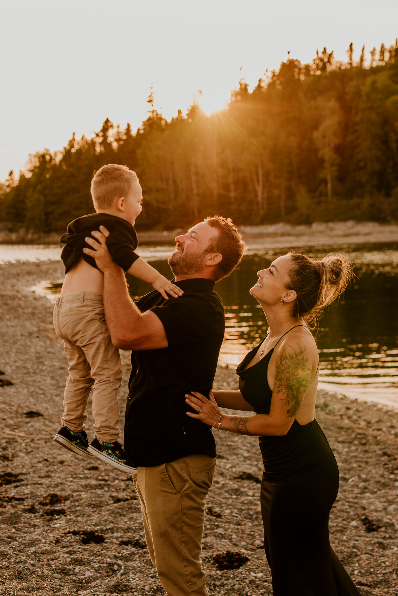 Family of three sharing a joyful moment at golden hour on a rocky beach, father lifting young son while mother smiles nearby.