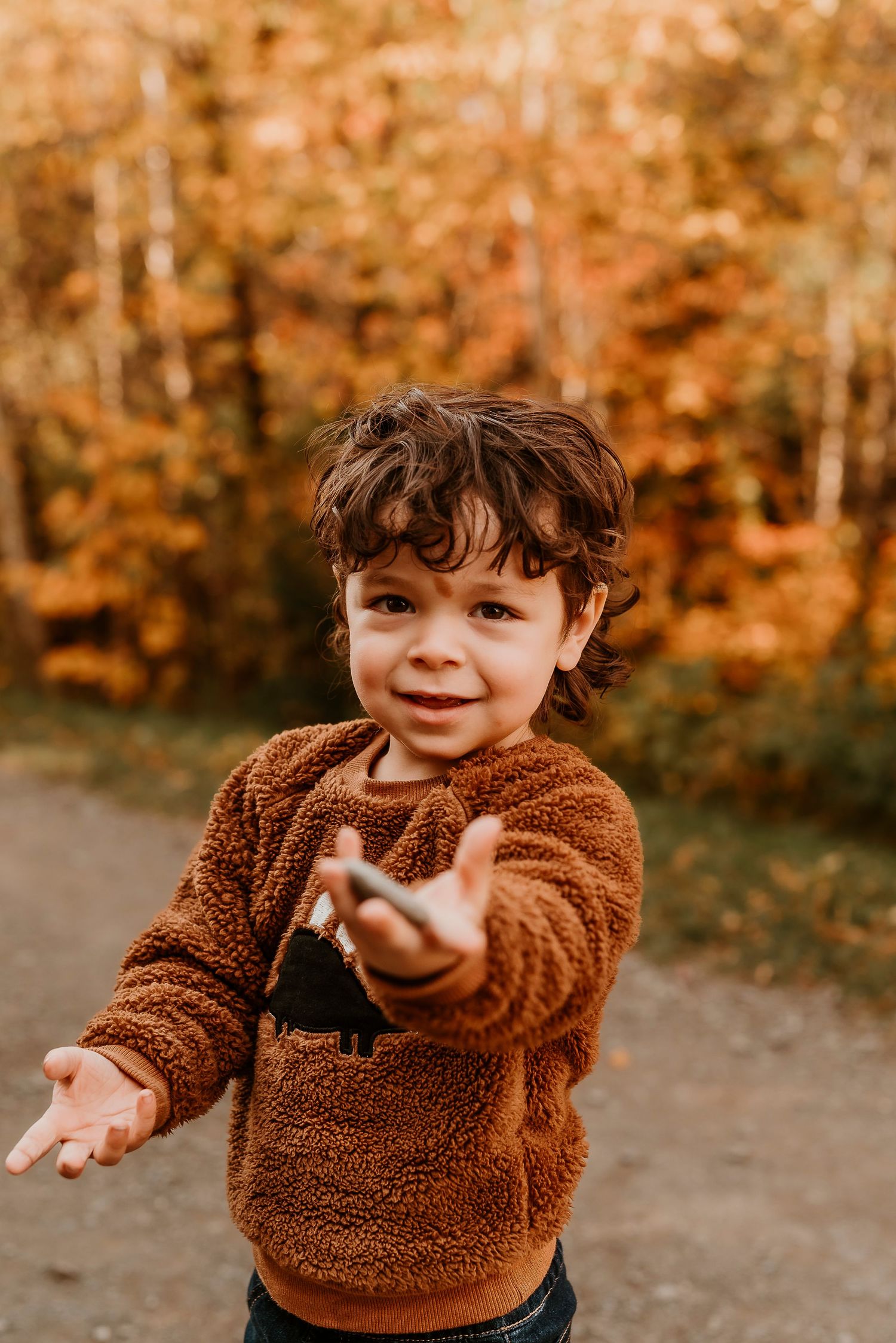A smiling toddler in a brown fuzzy sweater reaches toward the camera outdoors amid vibrant autumn foliage.