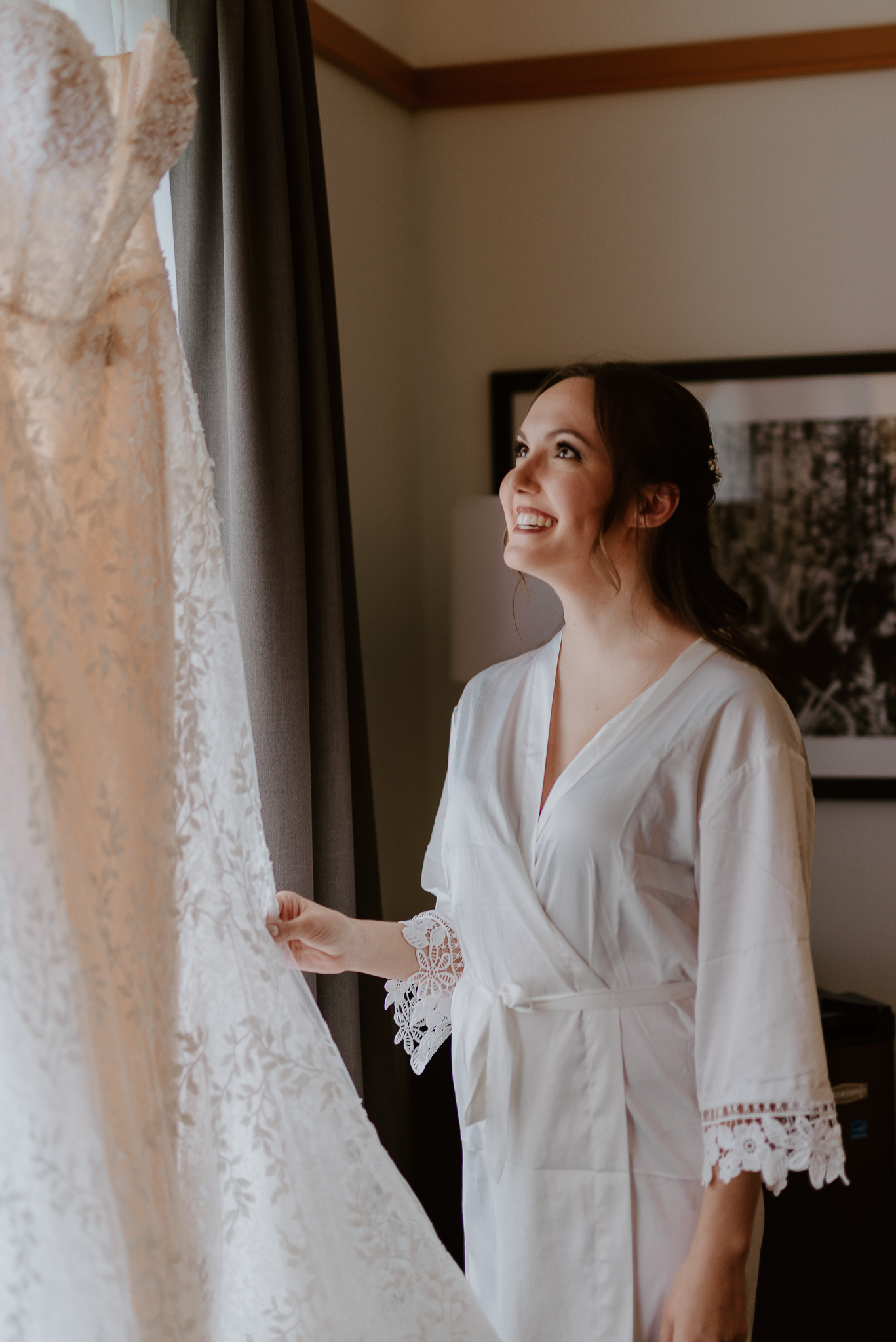A person in a white lace robe admires a wedding dress hanging by the window.
