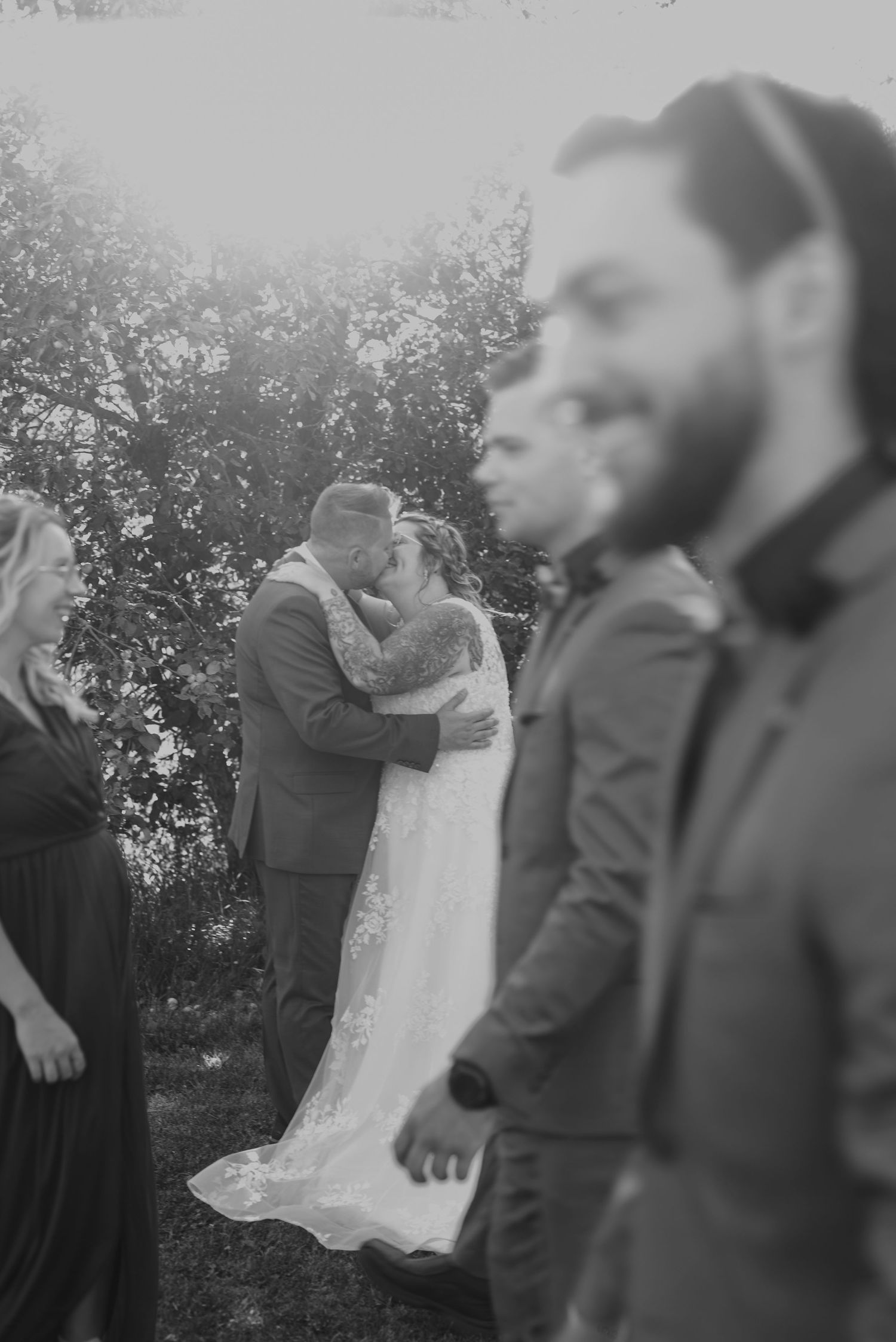 Une photo de mariage en noir et blanc d'un couple qui s'embrasse au loin; leurs invités d'honneur se promenant spontanément au premier plan.