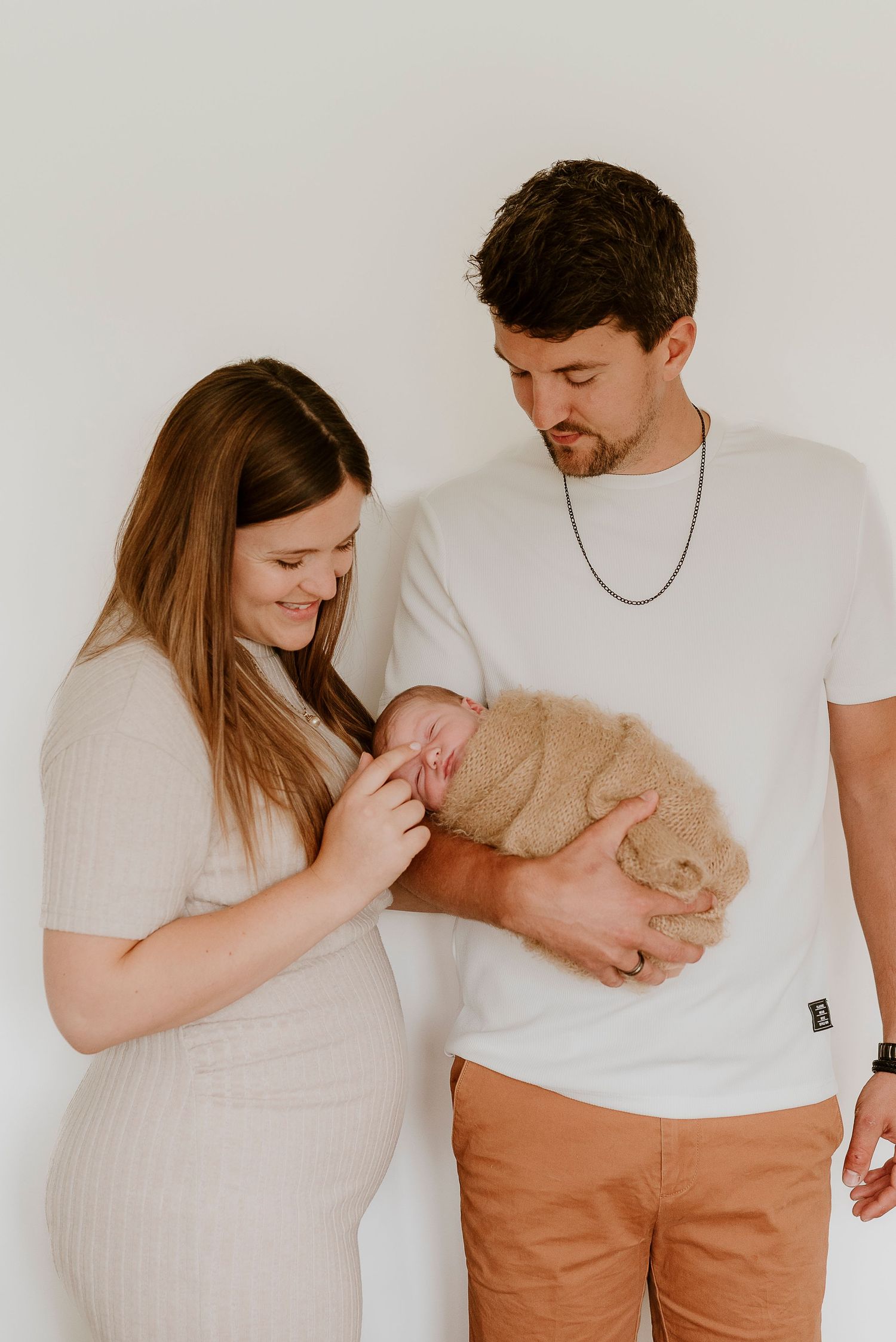 A young couple smiles lovingly at their newborn baby wrapped in a soft tan blanket during a newborn photo session.