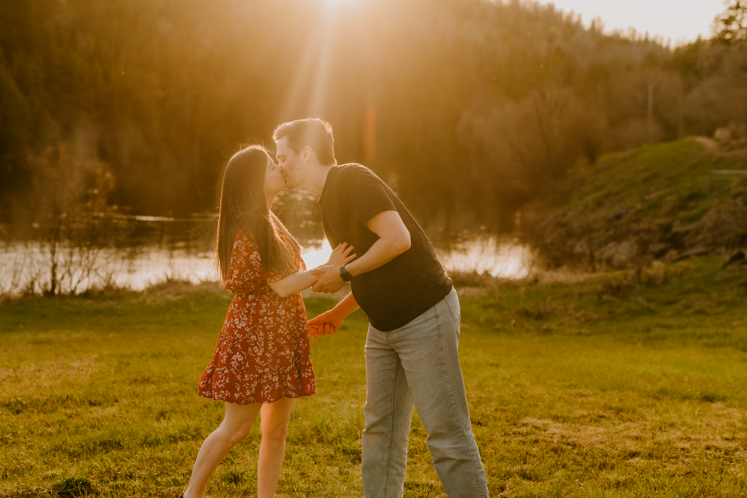 Couple dancing in golden sunset light by a lake, woman in red floral dress, man in black shirt and jeans.