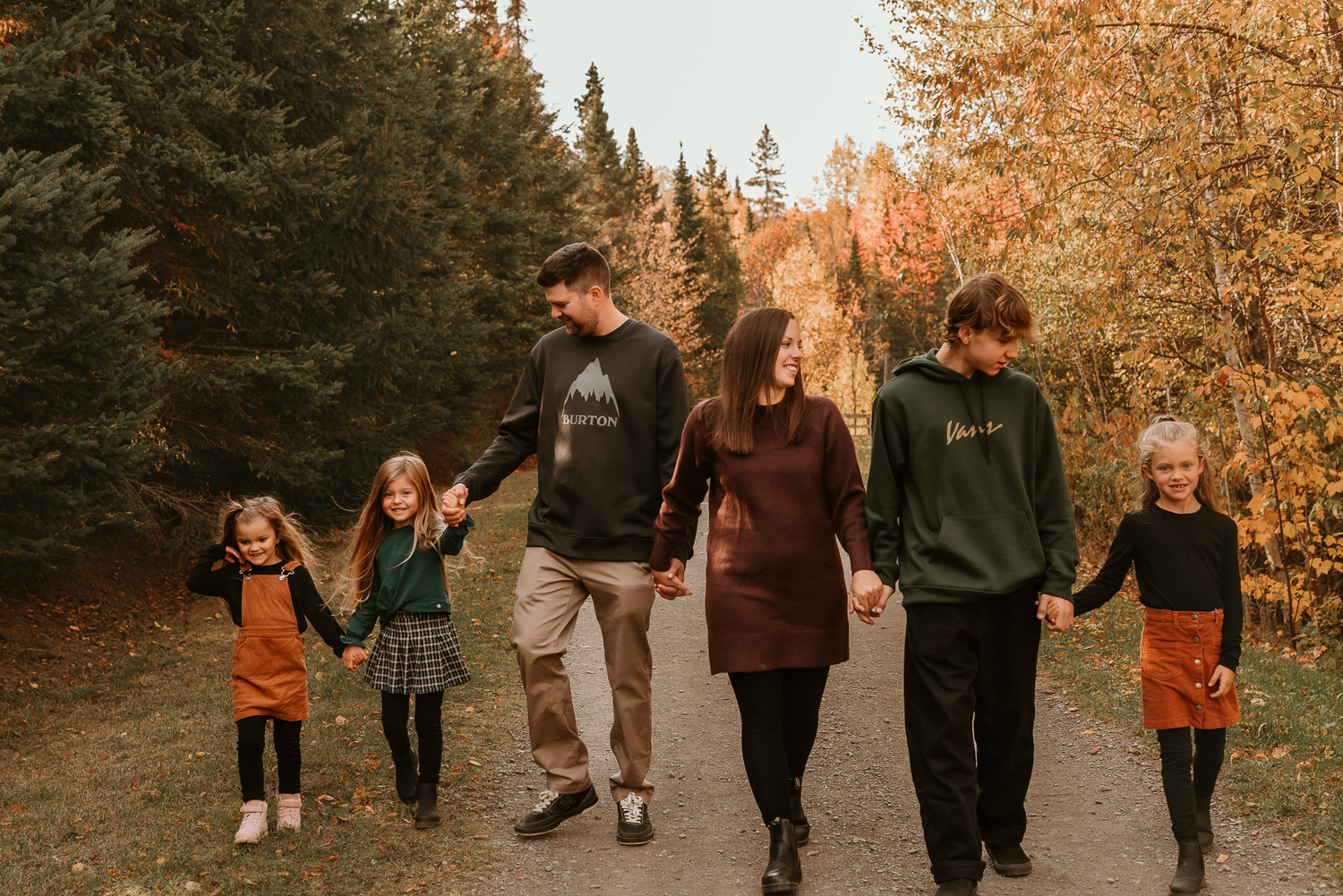 Family of five walking hand-in-hand on autumn forest path surrounded by colorful fall foliage.