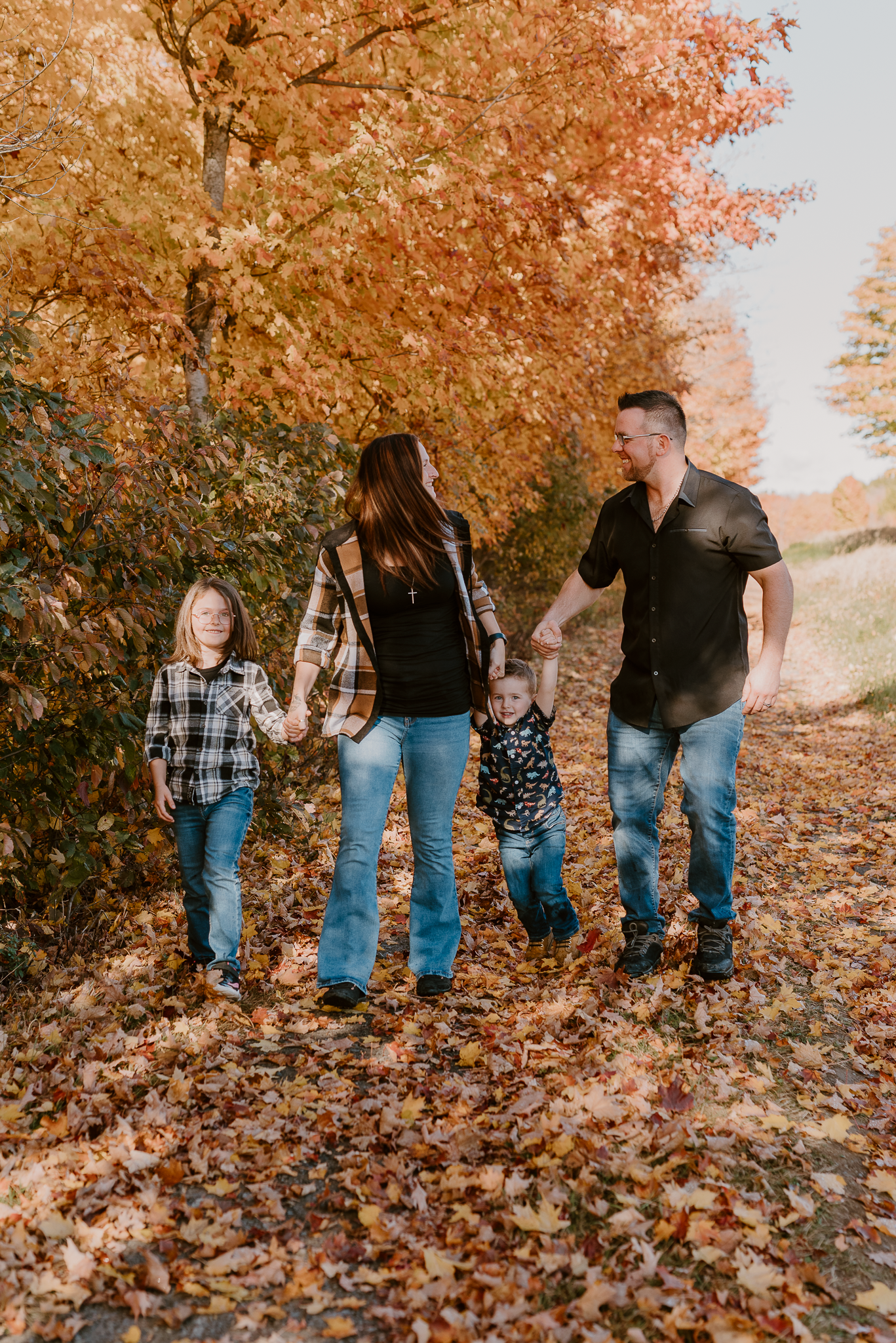 Family of four walking through autumn leaves on a tree-lined path, wearing flannel shirts and jeans.