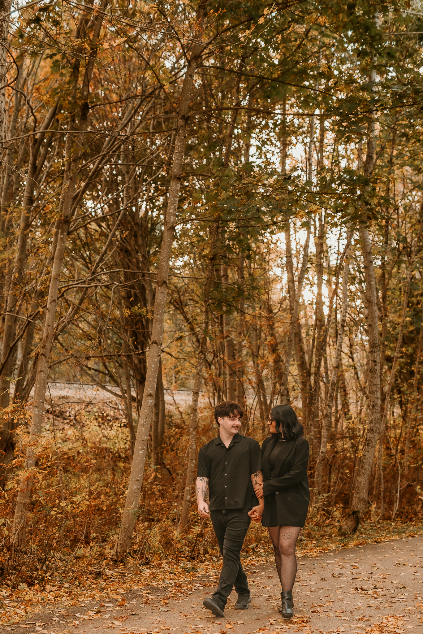 Couple holding hands walking on autumn forest path surrounded by golden fall foliage trees wearing matching black outfits.