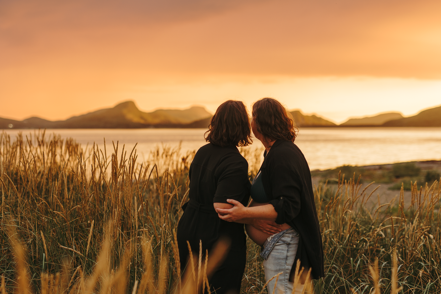 Couple embracing in golden wheat field at sunset with mountains and lake in background.