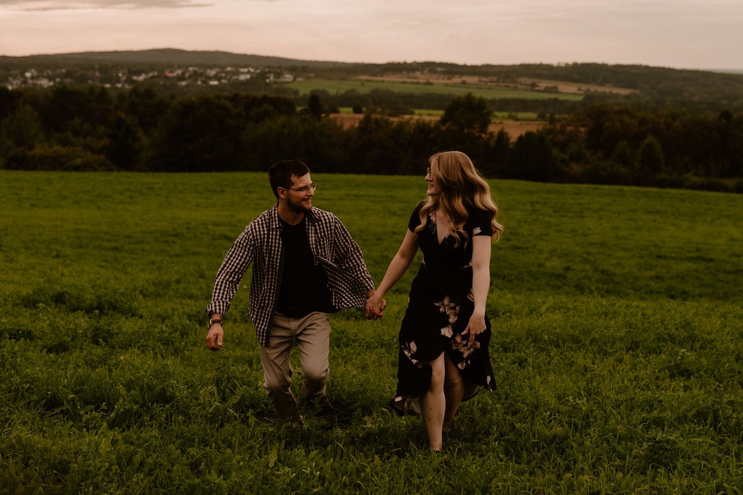 Romantic couple holding hands while walking through a lush green meadow at sunset with rolling hills in background.