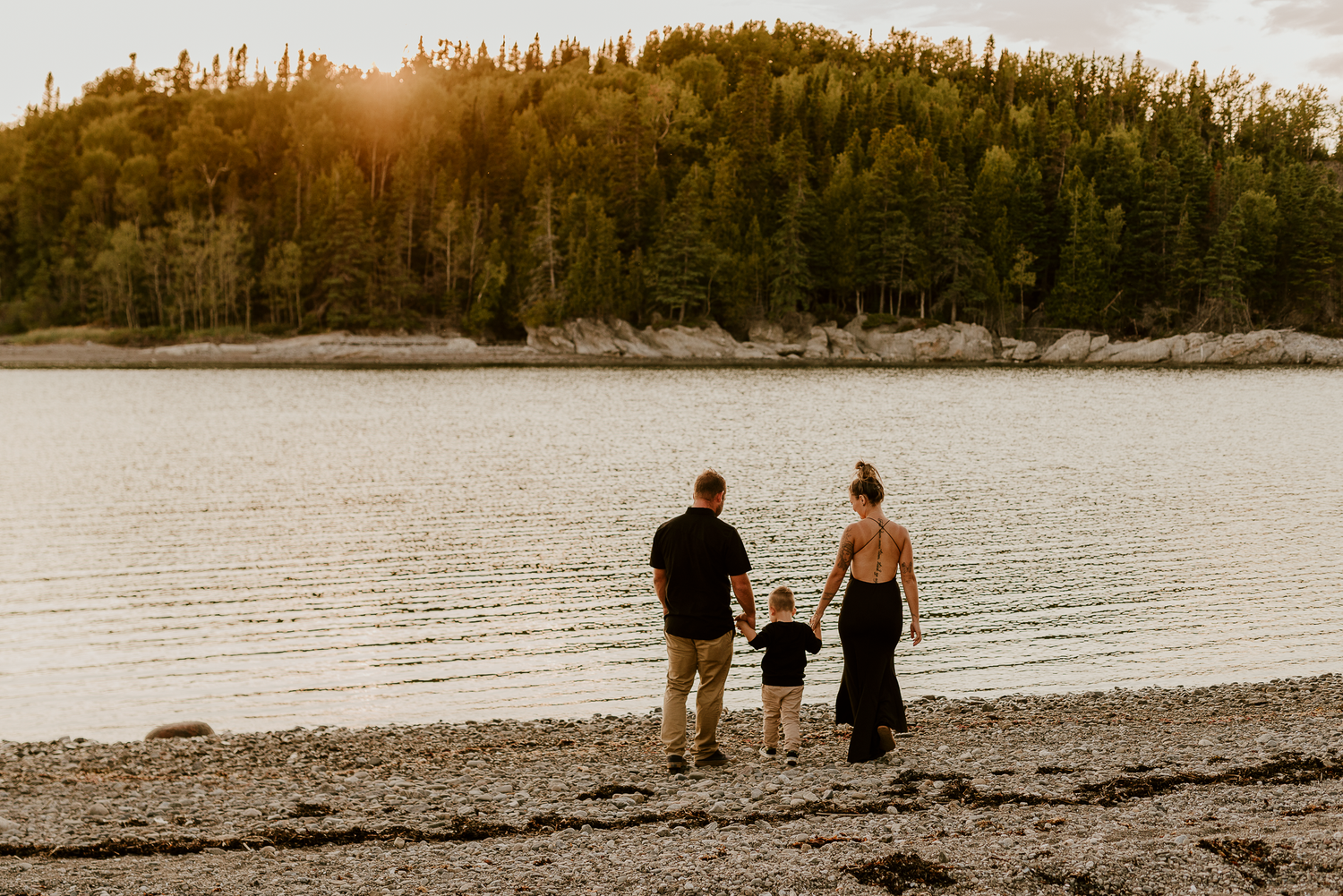 Family of three standing on rocky shoreline at golden hour, gazing at calm lake surrounded by forest.
