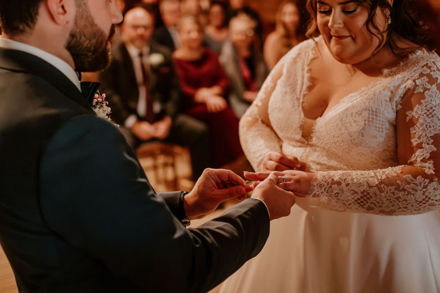 Bride and groom exchanging wedding rings during romantic ceremony with guests seated in background.