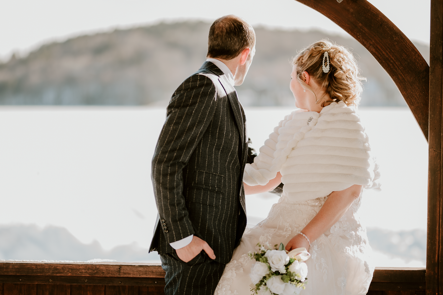 Bride and groom stand together overlooking scenic waterfront, bride holding white bouquet, groom in pinstripe suit.