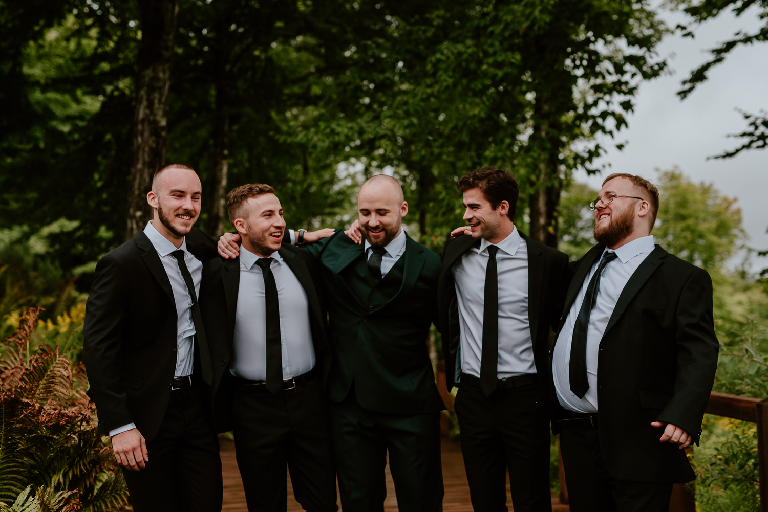 Groomsmen in black suits laugh together during outdoor wedding portraits.