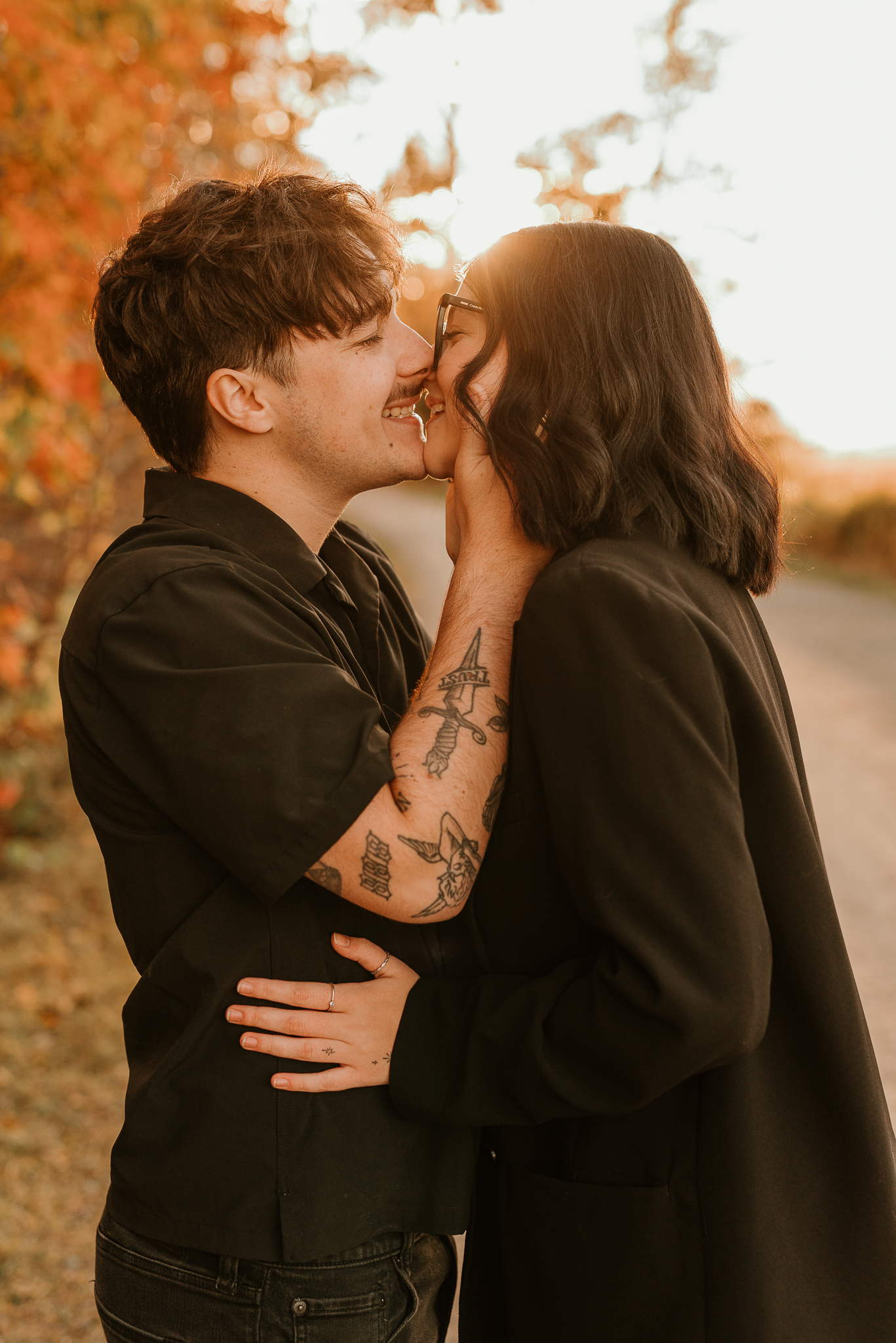 A couple in black outfits shares a romantic moment outdoors, surrounded by warm autumn foliage in golden light.