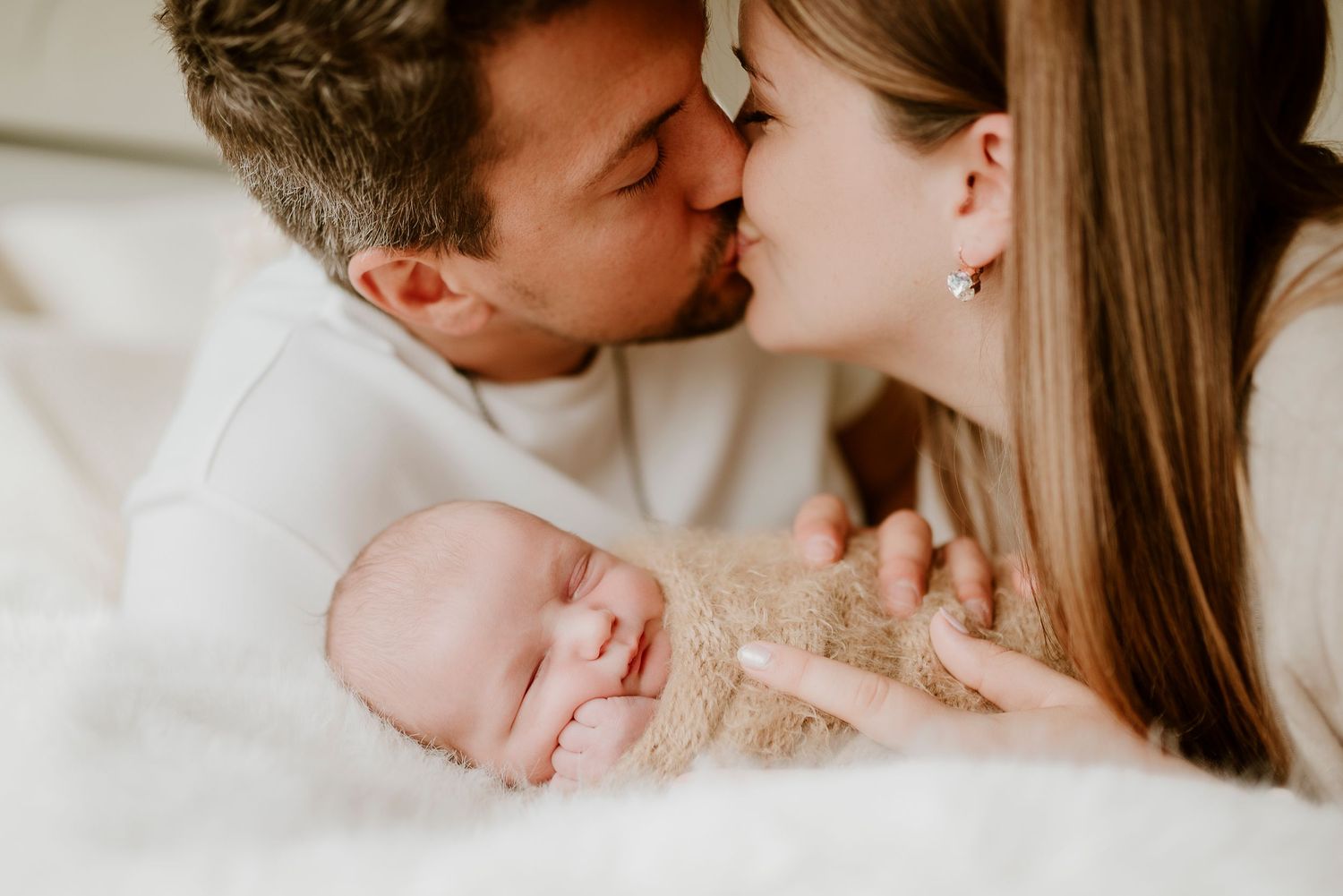 Parents sharing a kiss while cradling their sleeping newborn baby wrapped in a soft beige swaddle.