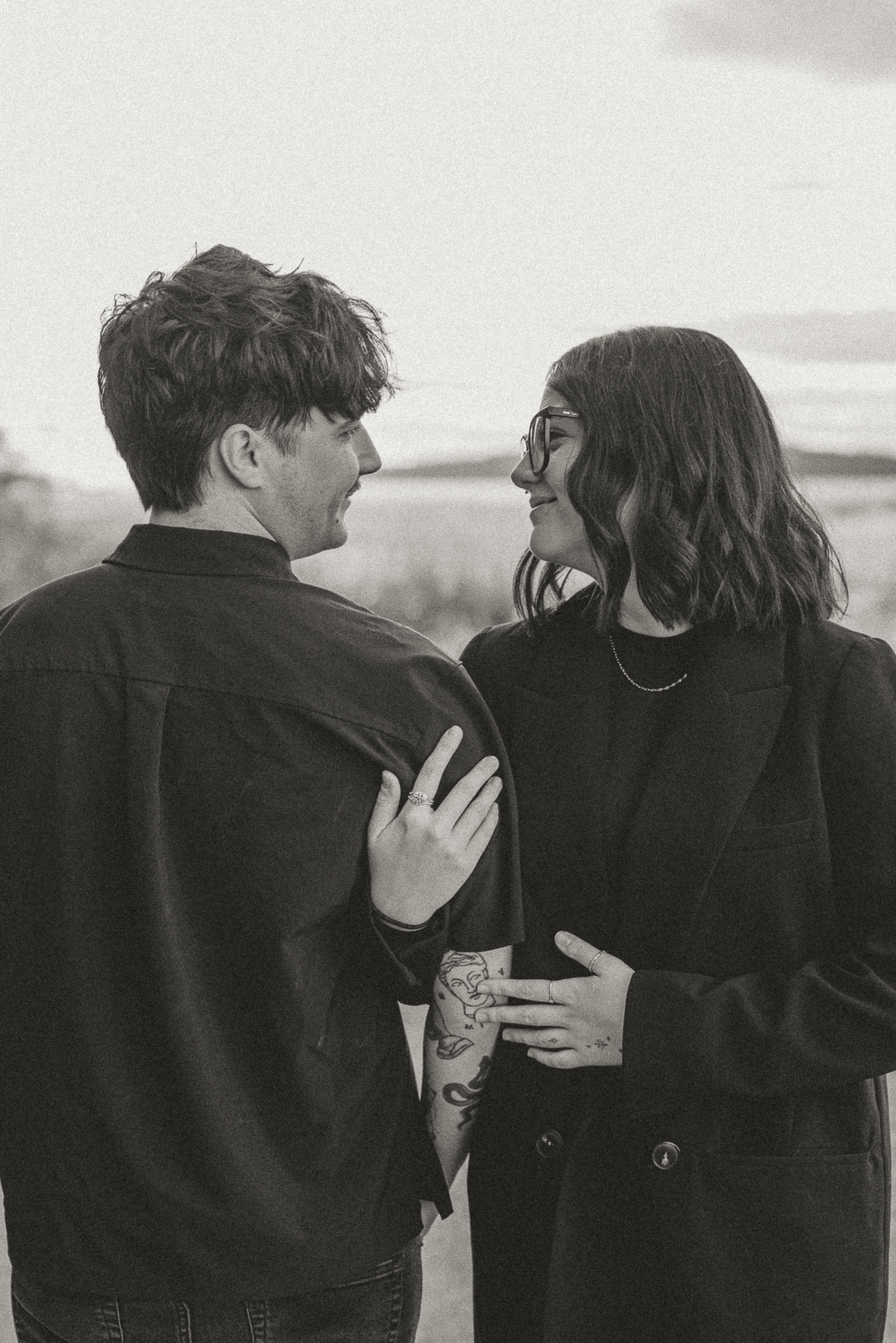 Black and white photo of a couple embracing outdoors, smiling at each other, woman wearing glasses and man with tattoos.
