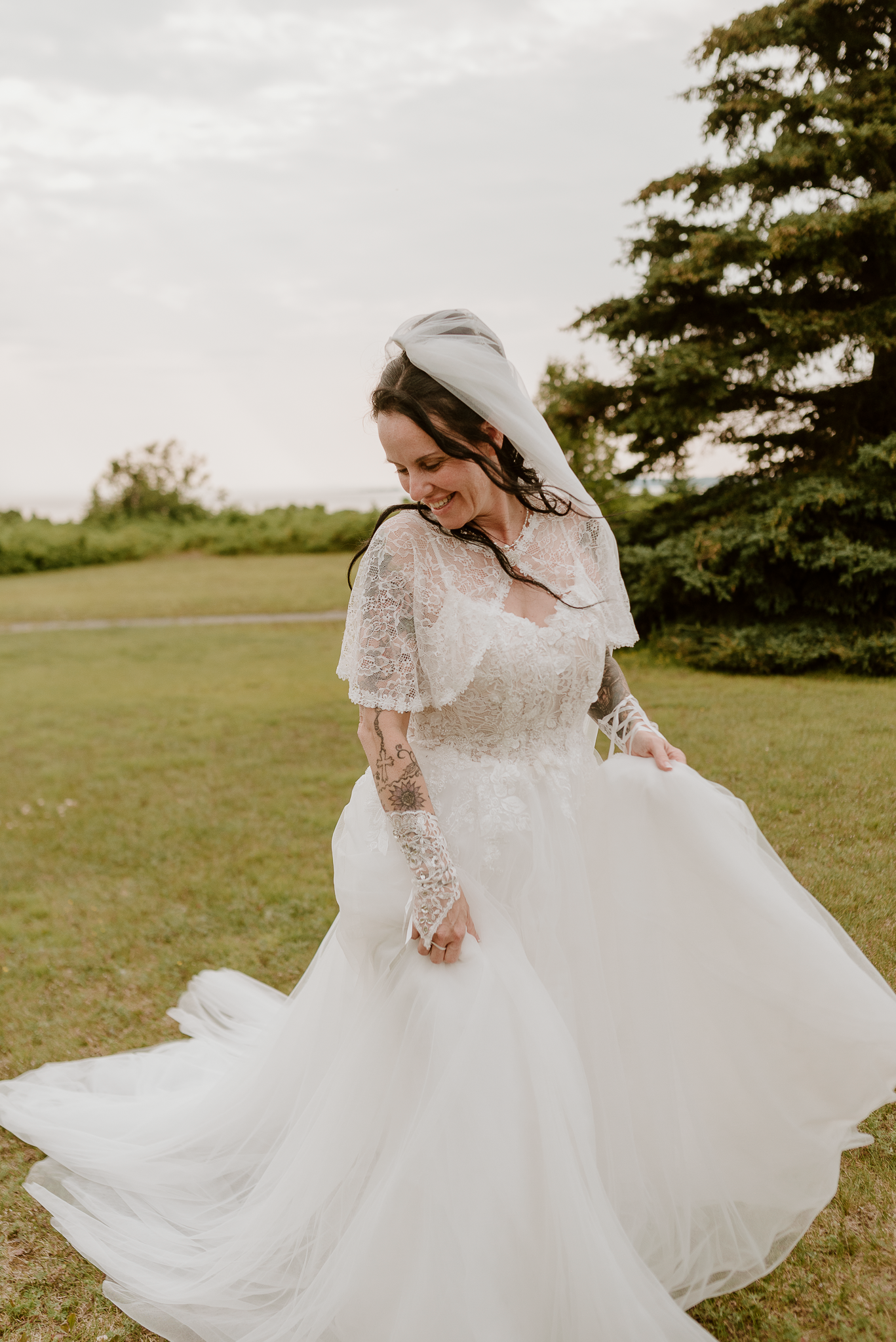 Bride in lace long-sleeve wedding gown and veil twirling on green lawn with trees in background.