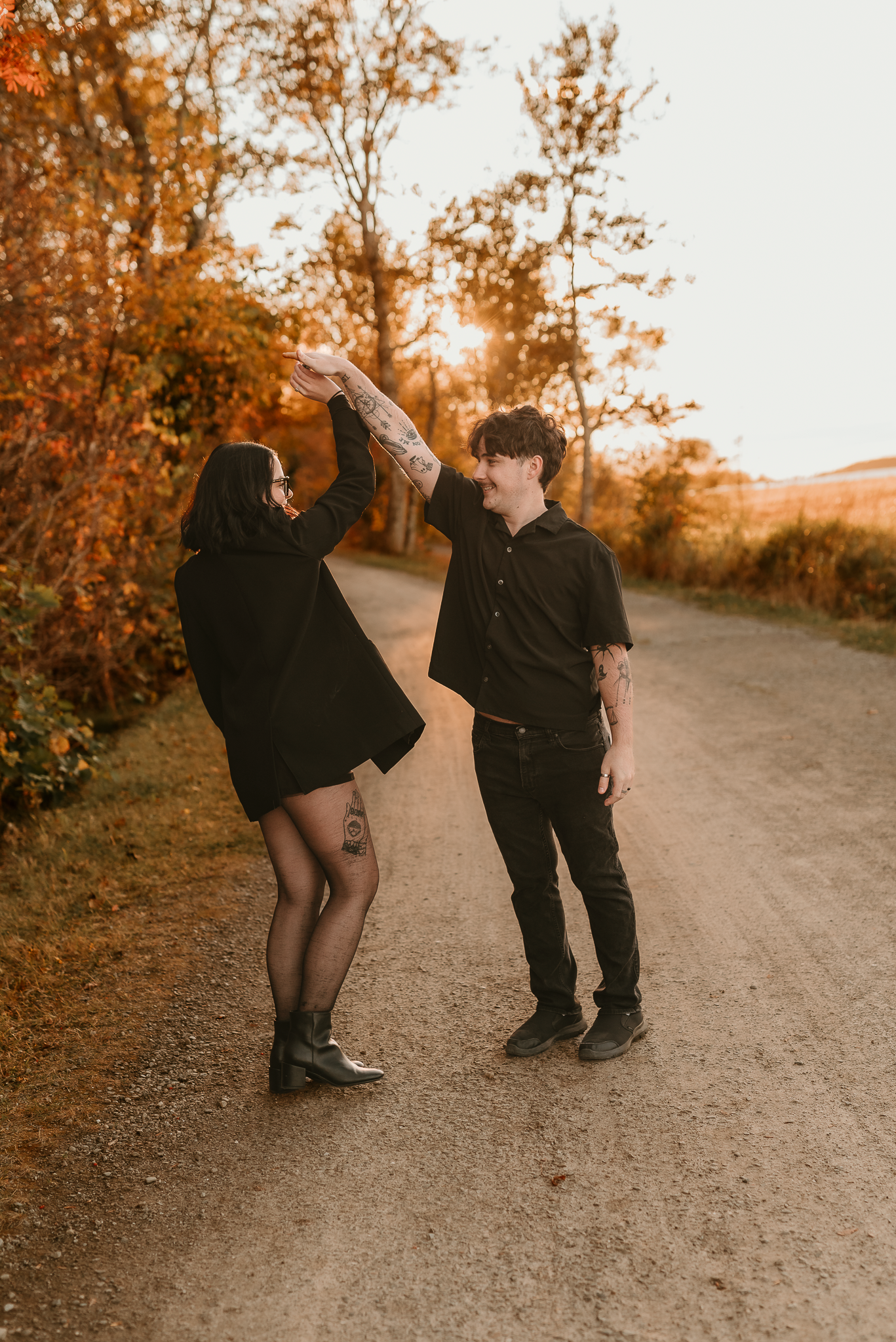Couple in matching black outfits dancing on a gravel path surrounded by autumn foliage at golden hour.