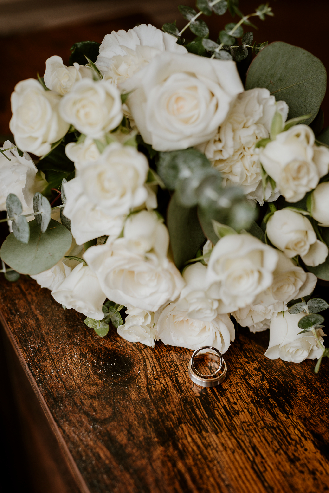 White roses and eucalyptus wedding bouquet with wedding rings on rustic wooden surface.
