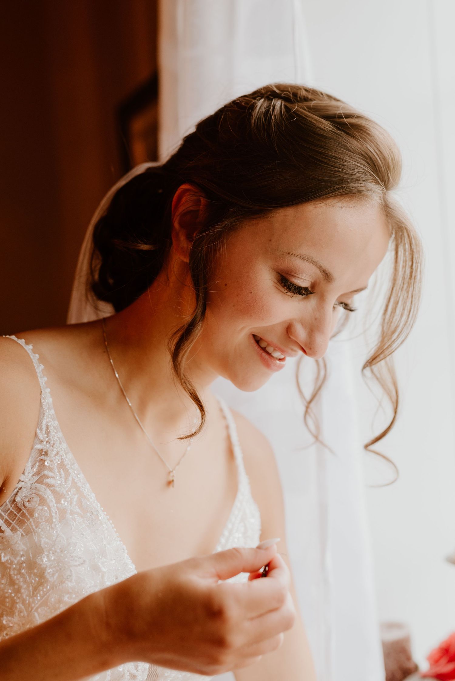 A bride in a lace dress smiles softly while getting ready by a window.