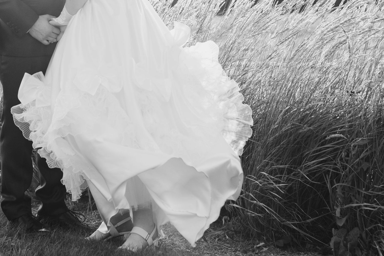 La délicate crinoline d'une mariée qui tient les mains de son époux devant un champ d'herbes hautes, le tout capté en noir et blanc.