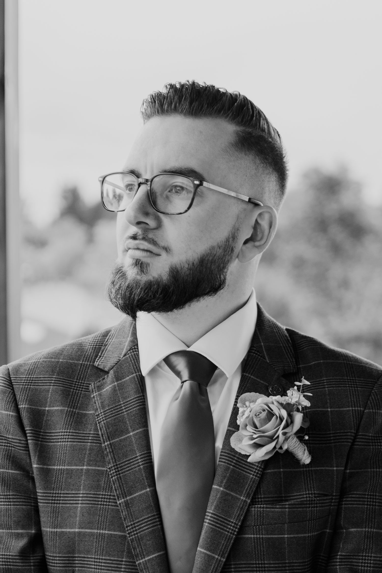 Groom in plaid suit jacket and glasses poses with boutonniere for wedding portrait.