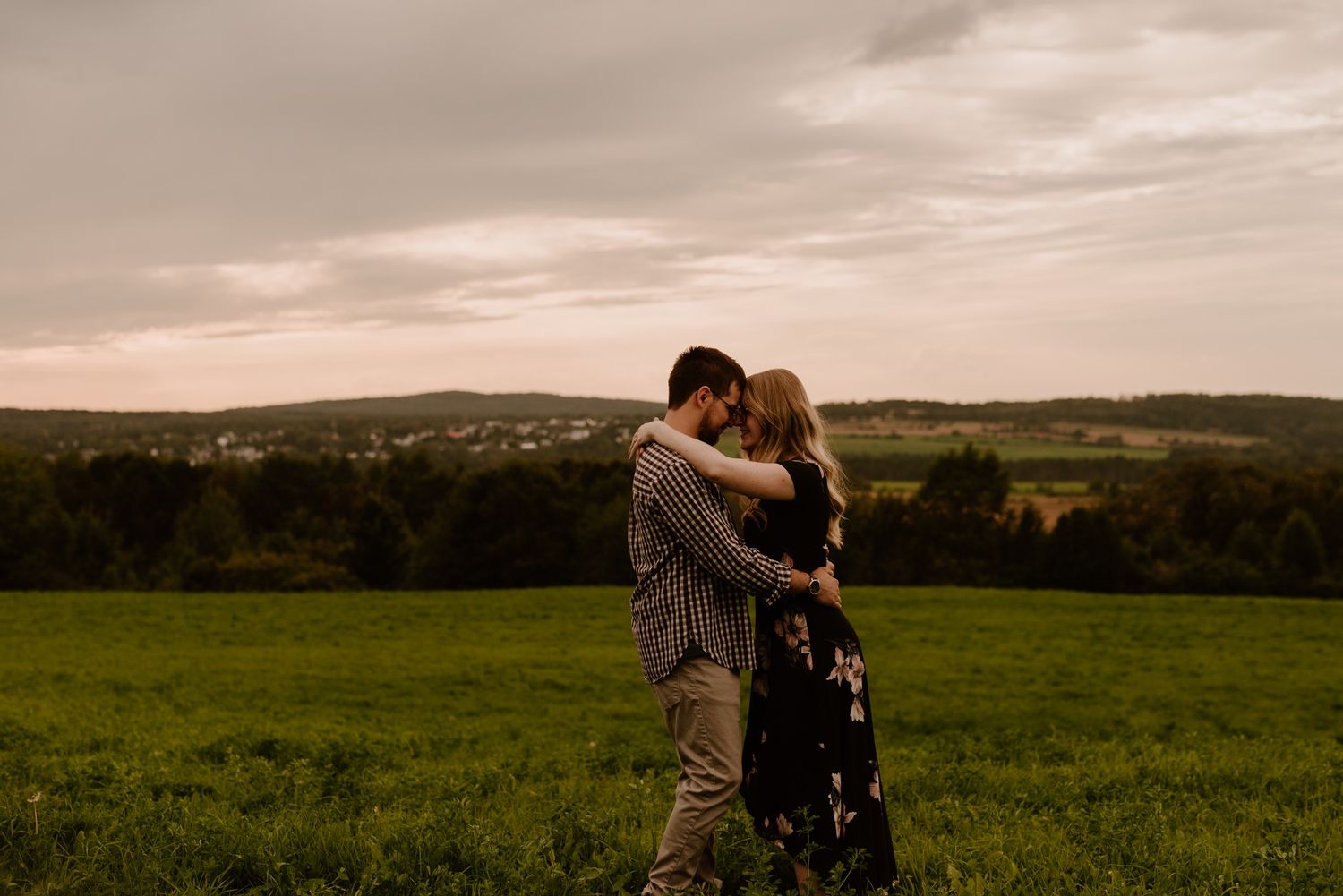 Couple embracing in field at sunset with rolling hills in background.