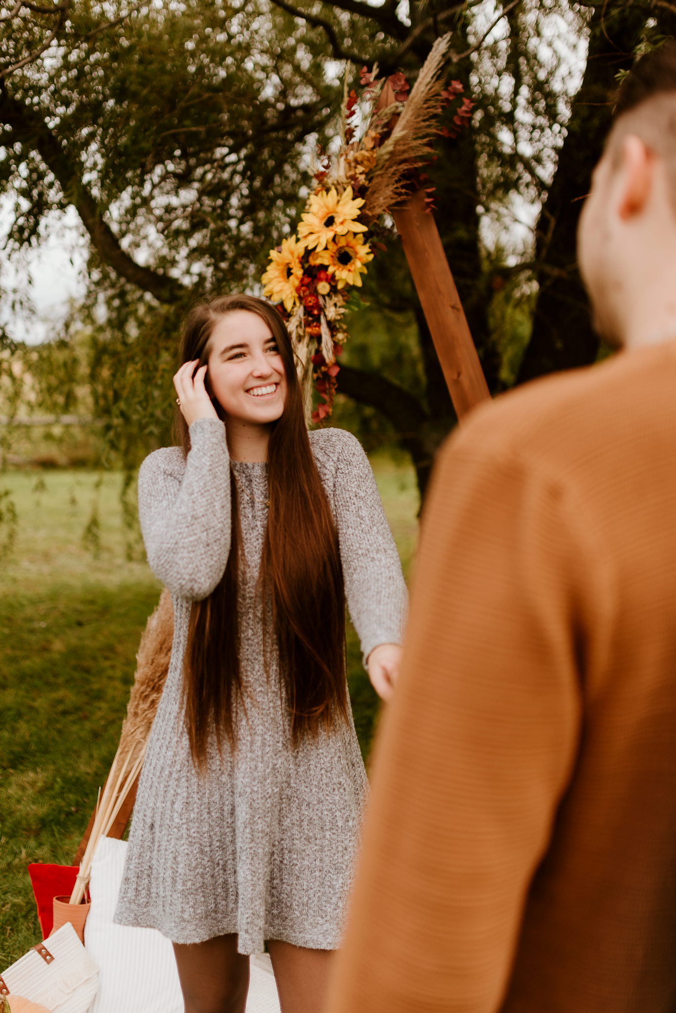 A smiling young woman with long brown hair in a gray dress stands outdoors near a floral arch with sunflowers.
