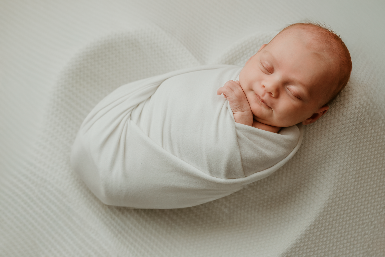 Peaceful newborn baby swaddled in white wrap sleeping on soft white blanket in professional studio photo.