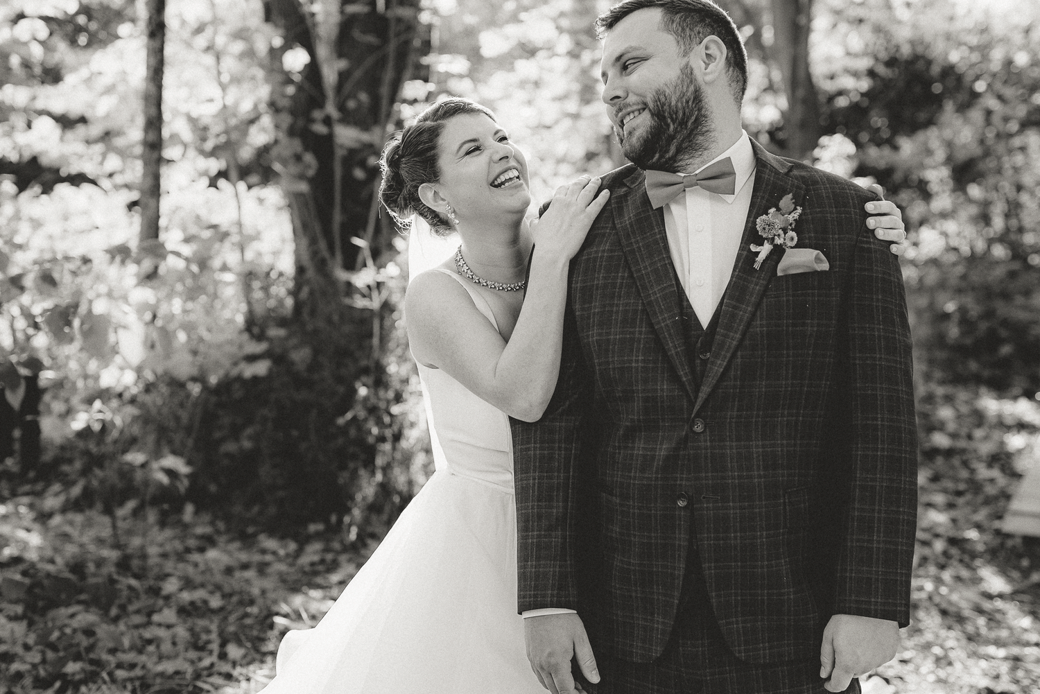 Black and white wedding photo of a laughing bride and bearded groom in plaid suit with bow tie outdoors.