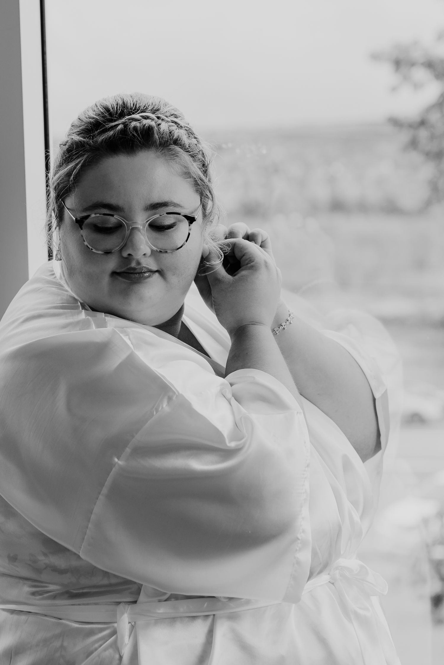 Black and white portrait of a bride in glasses and white robe getting ready for her wedding day.