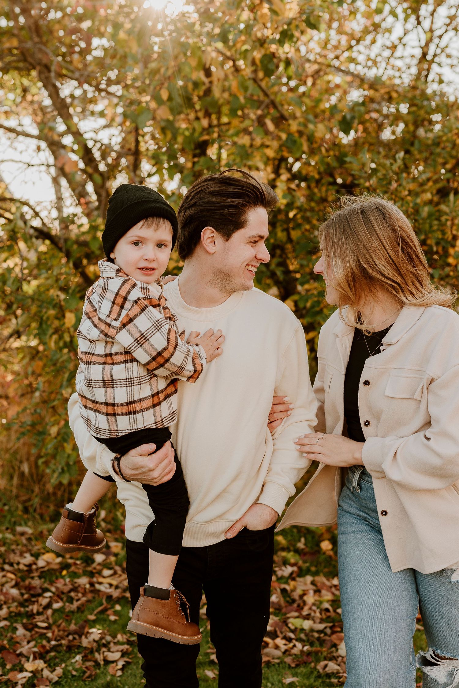 A young family poses outdoors in autumn, father holding toddler in plaid shirt while mother smiles warmly beside them.
