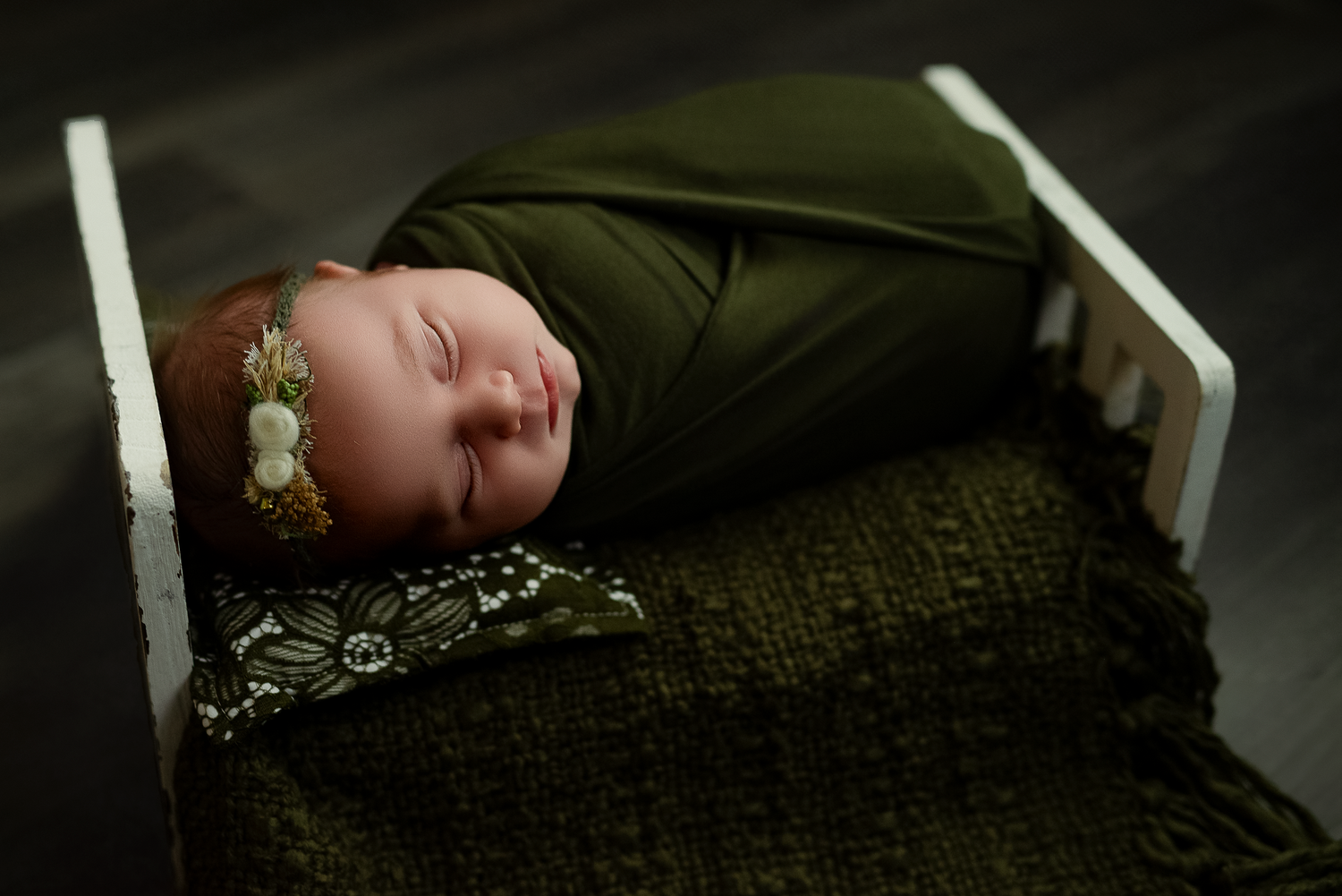Newborn baby swaddled in dark green wrap with floral headband, sleeping peacefully in a white wooden mini bed.