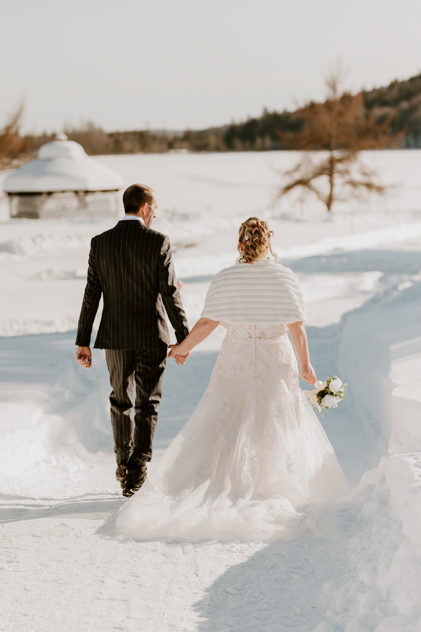 Bride and groom walking hand in hand through snowy winter landscape, bride holding bouquet in white gown with cape.