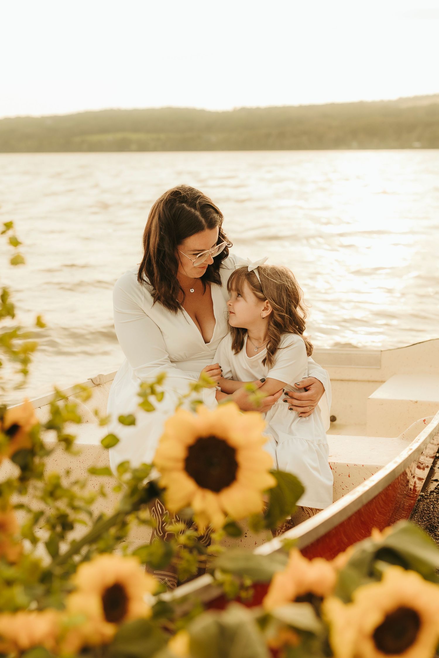 A mother and daughter in white dresses share a tender moment in a boat, surrounded by sunflowers with a lake backdrop.