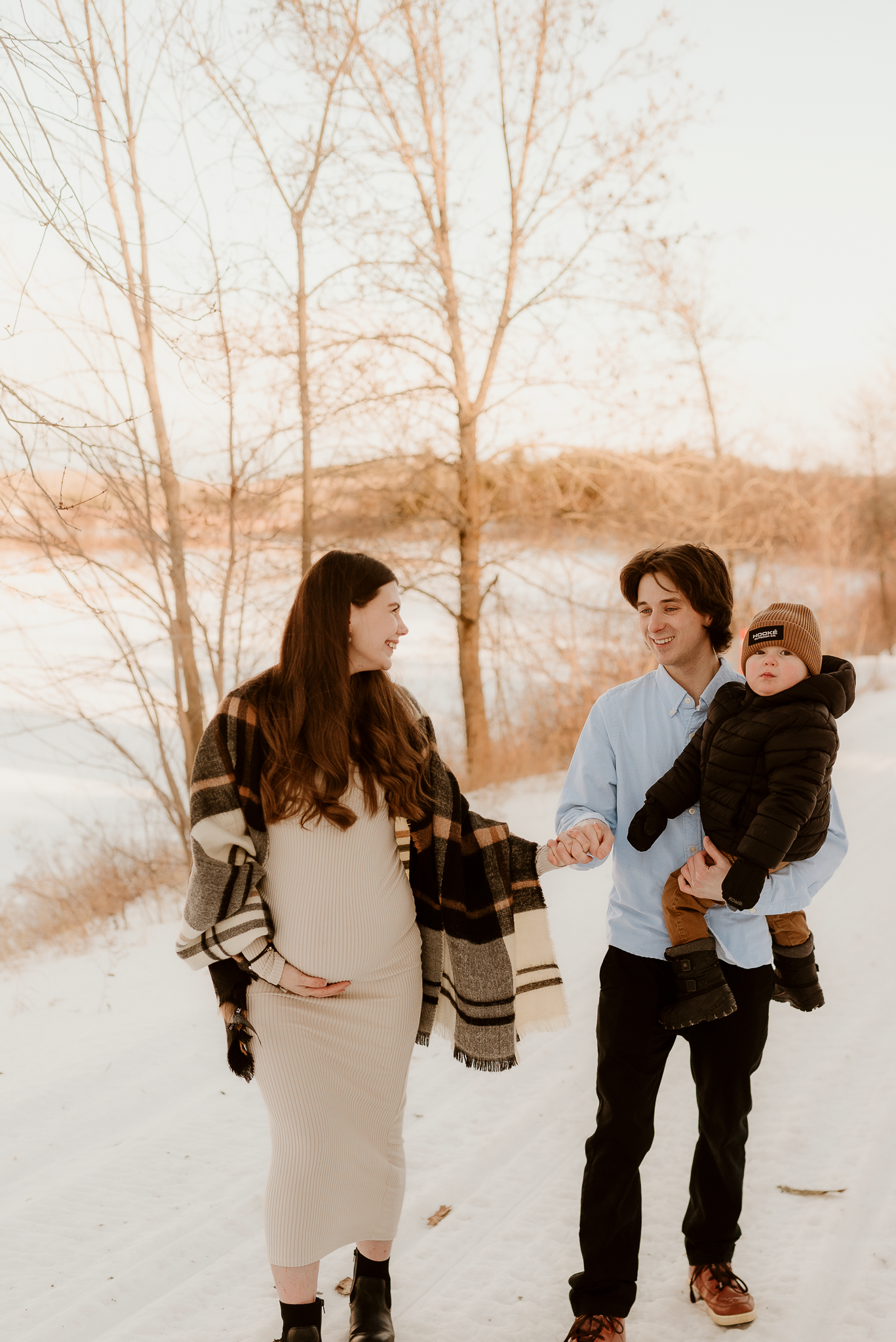 Pregnant woman in plaid shawl holds hands with man carrying toddler during winter maternity photo session in snowy landscape.