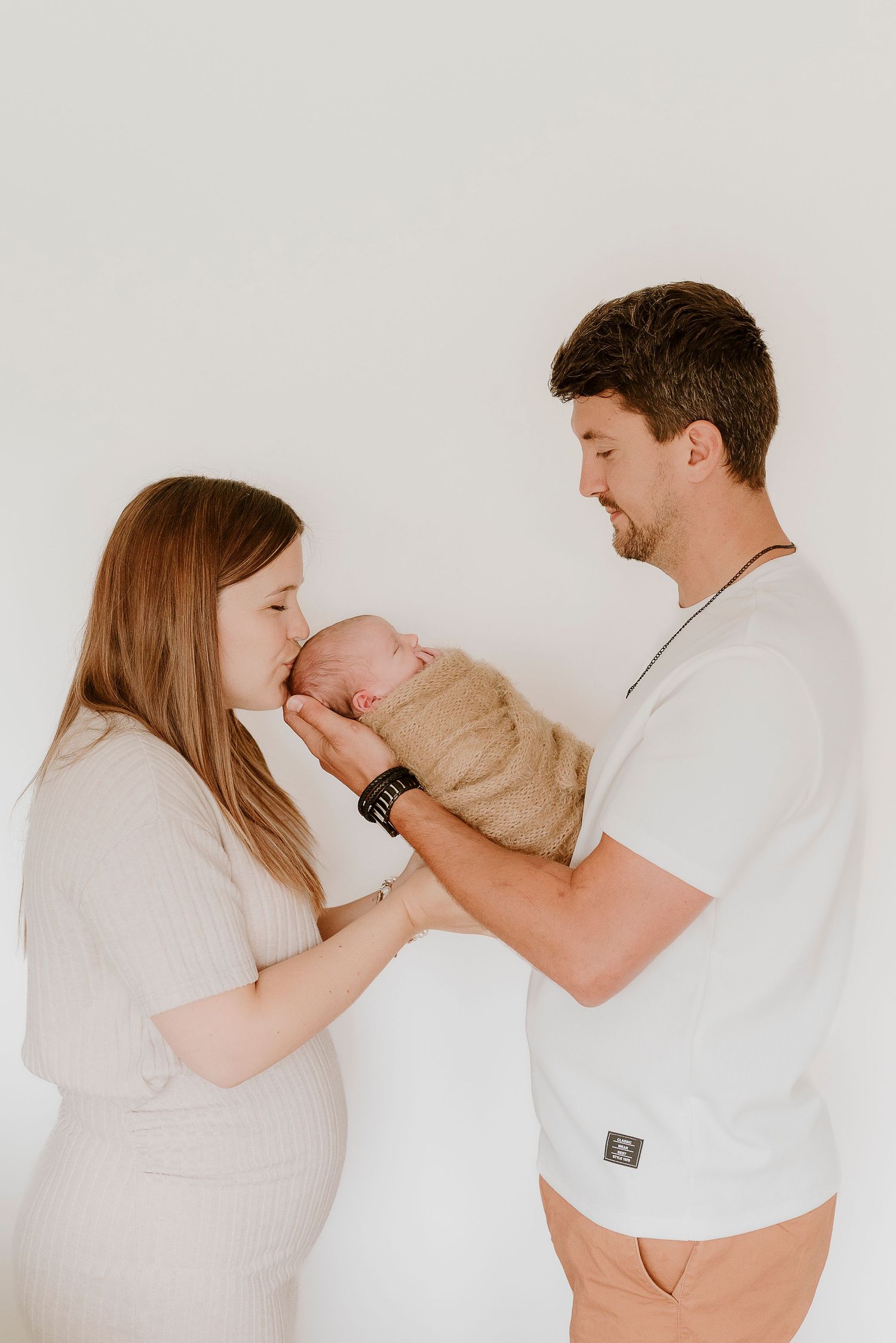 New parents smiling at their sleeping newborn baby wrapped in a tan swaddle blanket.