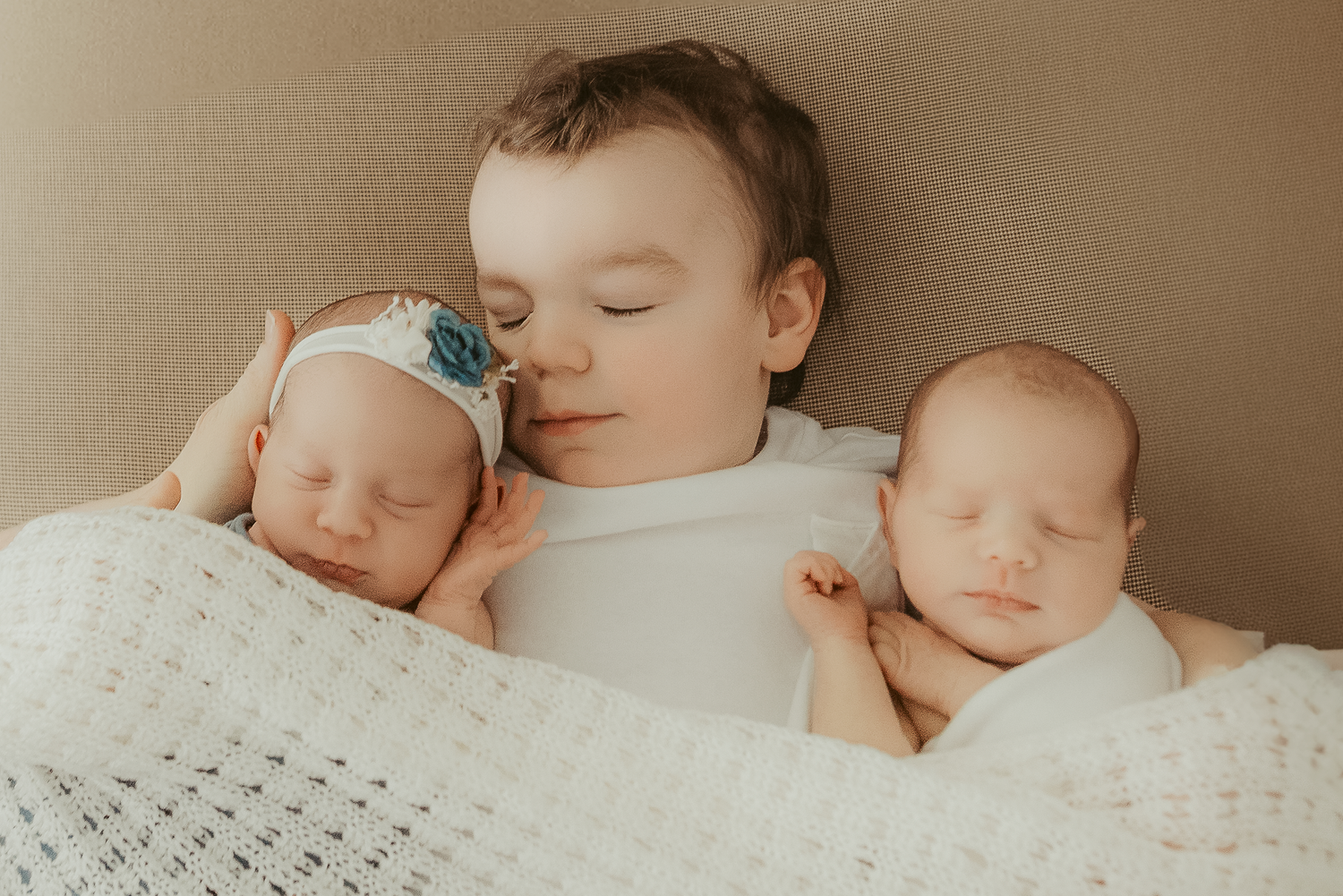 Sleeping toddler boy cuddles twin newborn babies wrapped in white blankets on a beige background.