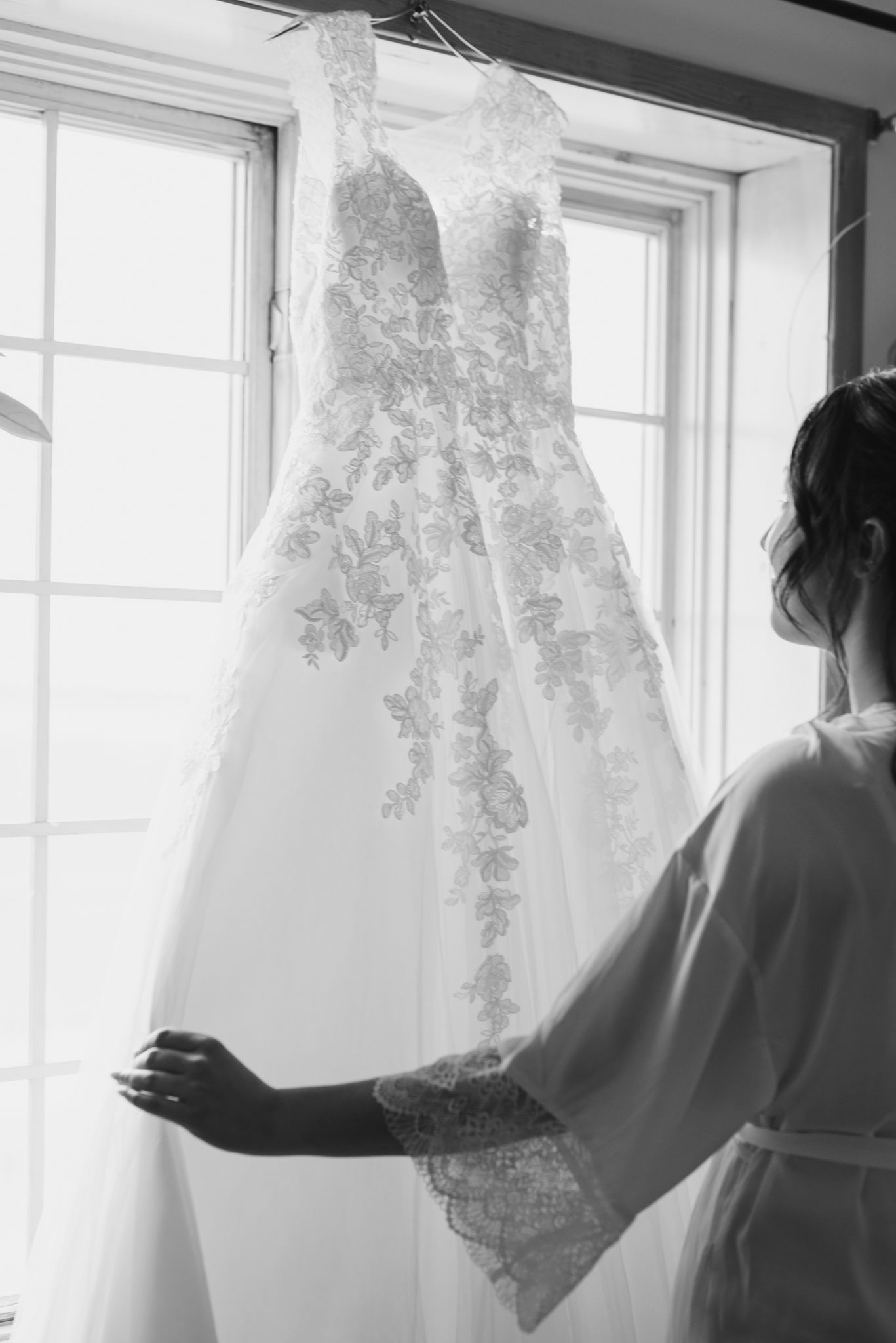 Admirative, une future mariée contemple sa robe suspendue devant des fenêtres très ensoleillées, le tout capté en noir et blanc.