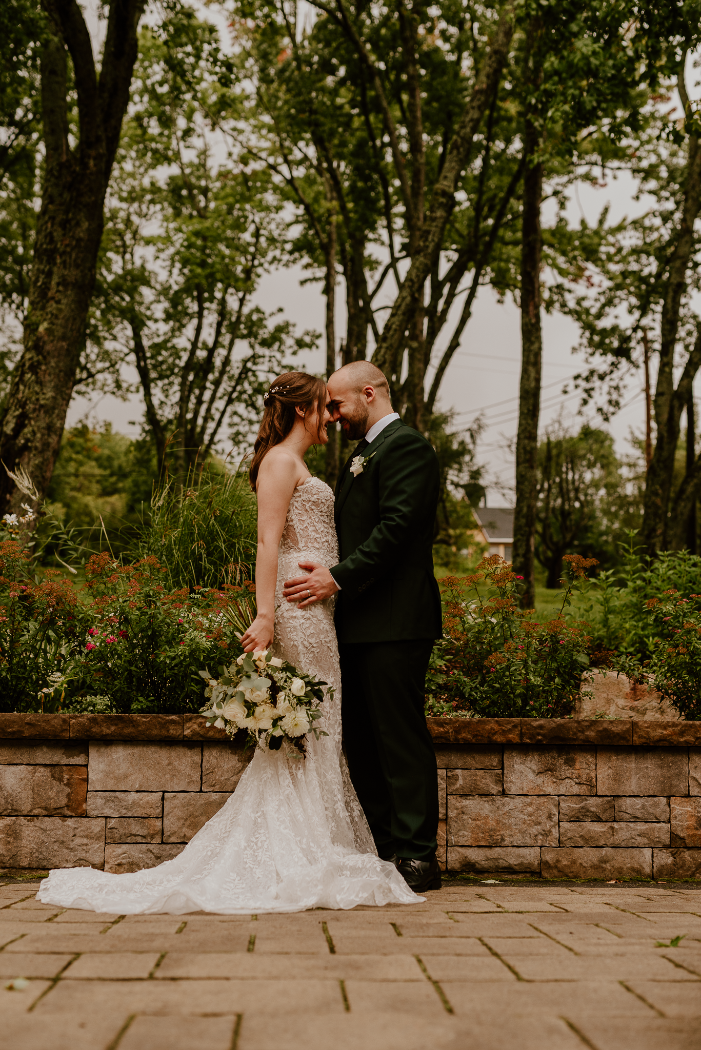 Bride and groom share an intimate moment in a garden setting with stone walls.