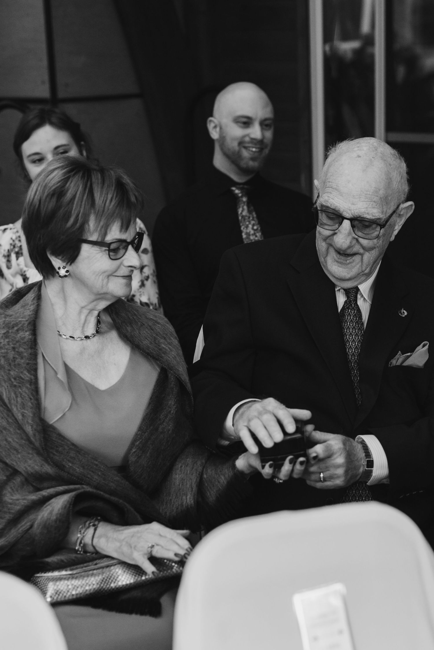 Wedding guests smile while looking at something on phone screen.