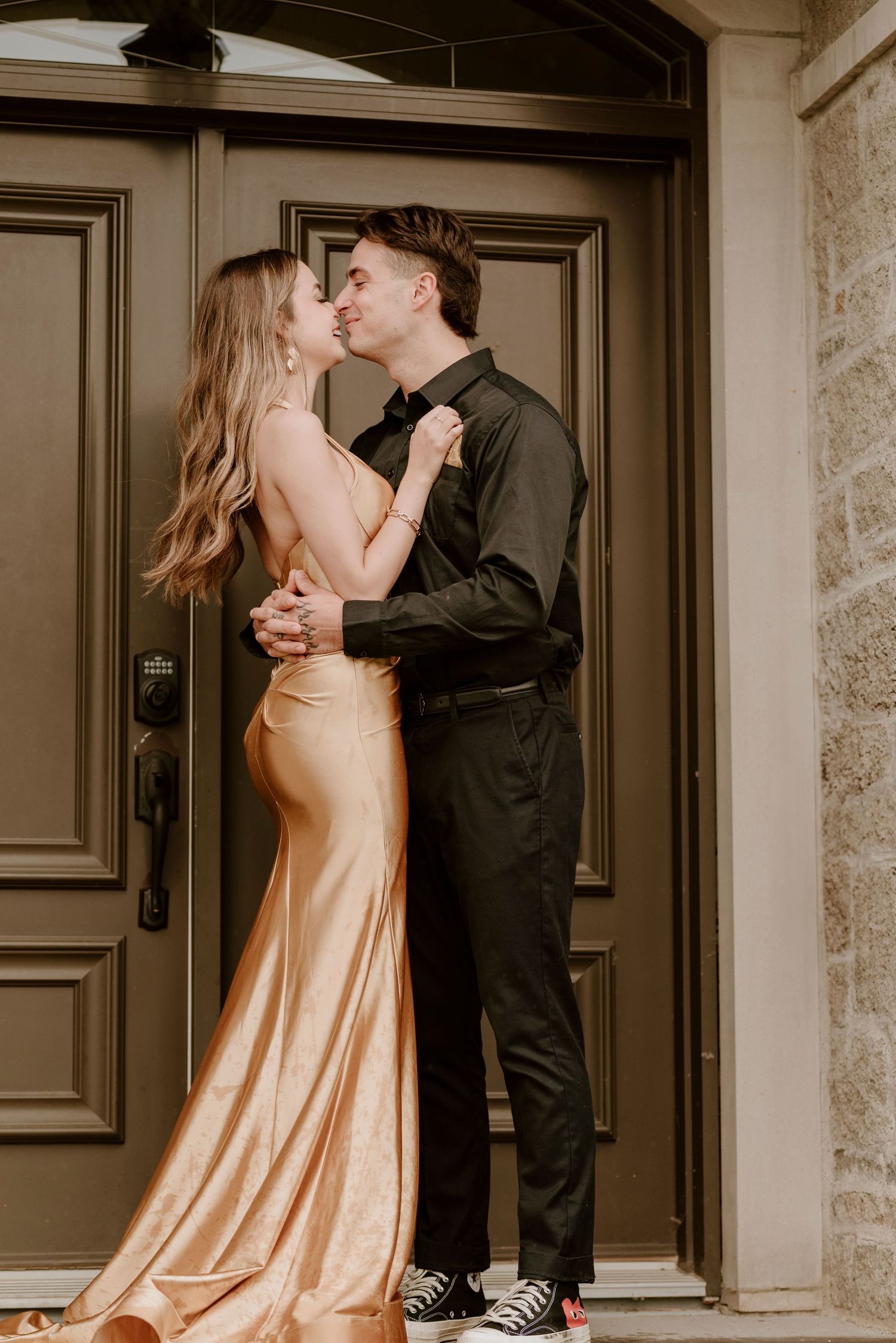 Couple in formal attire kissing in front of elegant door, woman in gold satin gown, man in black suit.