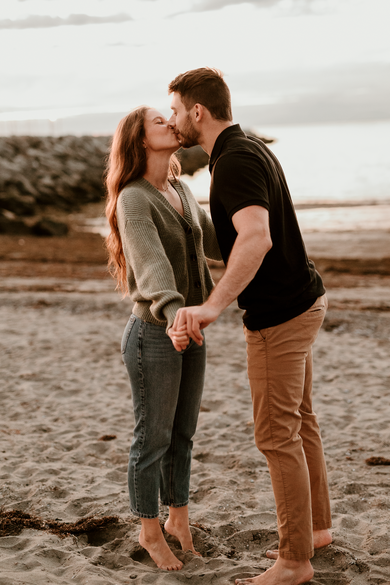 A couple shares a romantic kiss on a sandy beach at sunset, holding hands barefoot in casual outfits.