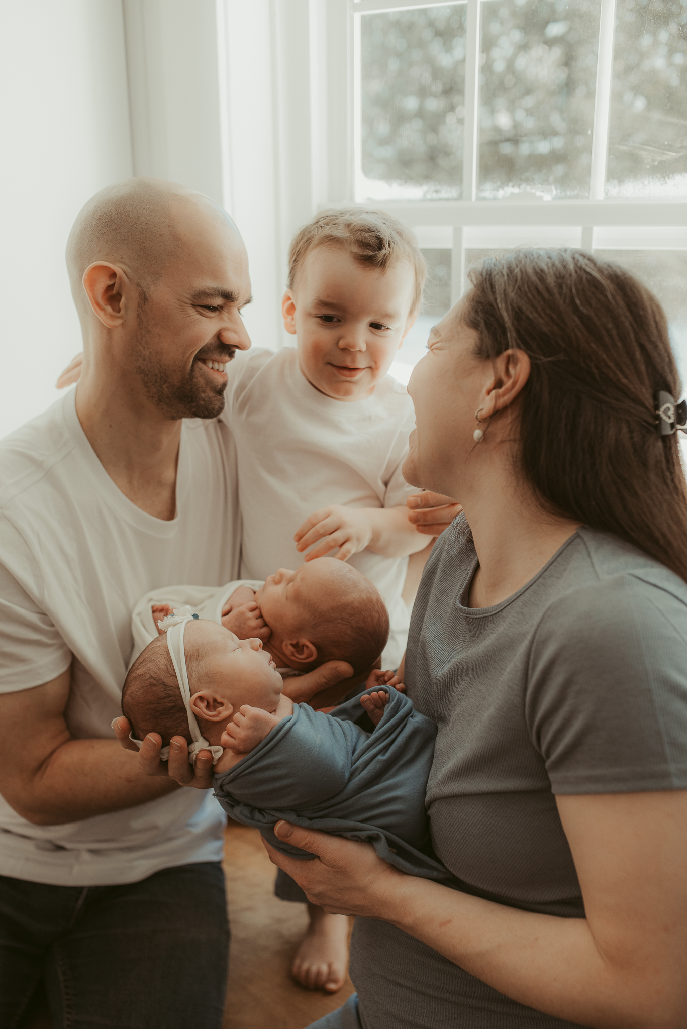 A happy family of four with parents holding newborn twins while their toddler smiles near a bright window.