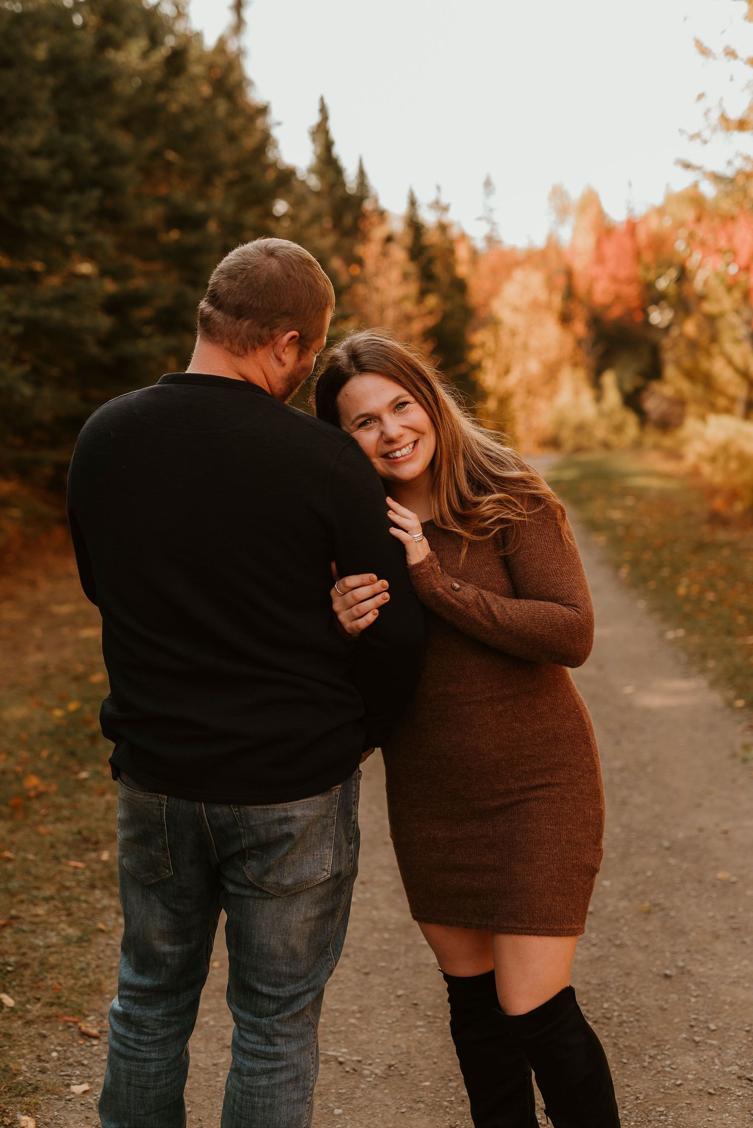 A smiling woman in a brown dress and black boots embraces a man on an autumn trail surrounded by colorful fall foliage.