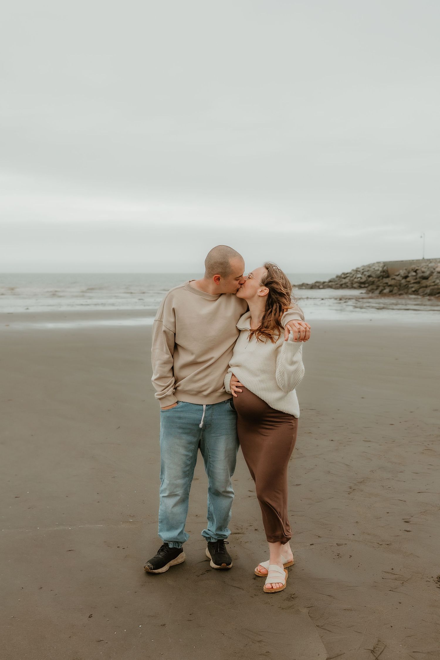Couple sharing a tender kiss on a sandy beach, woman in brown skirt and white sweater, man in jeans and beige top.