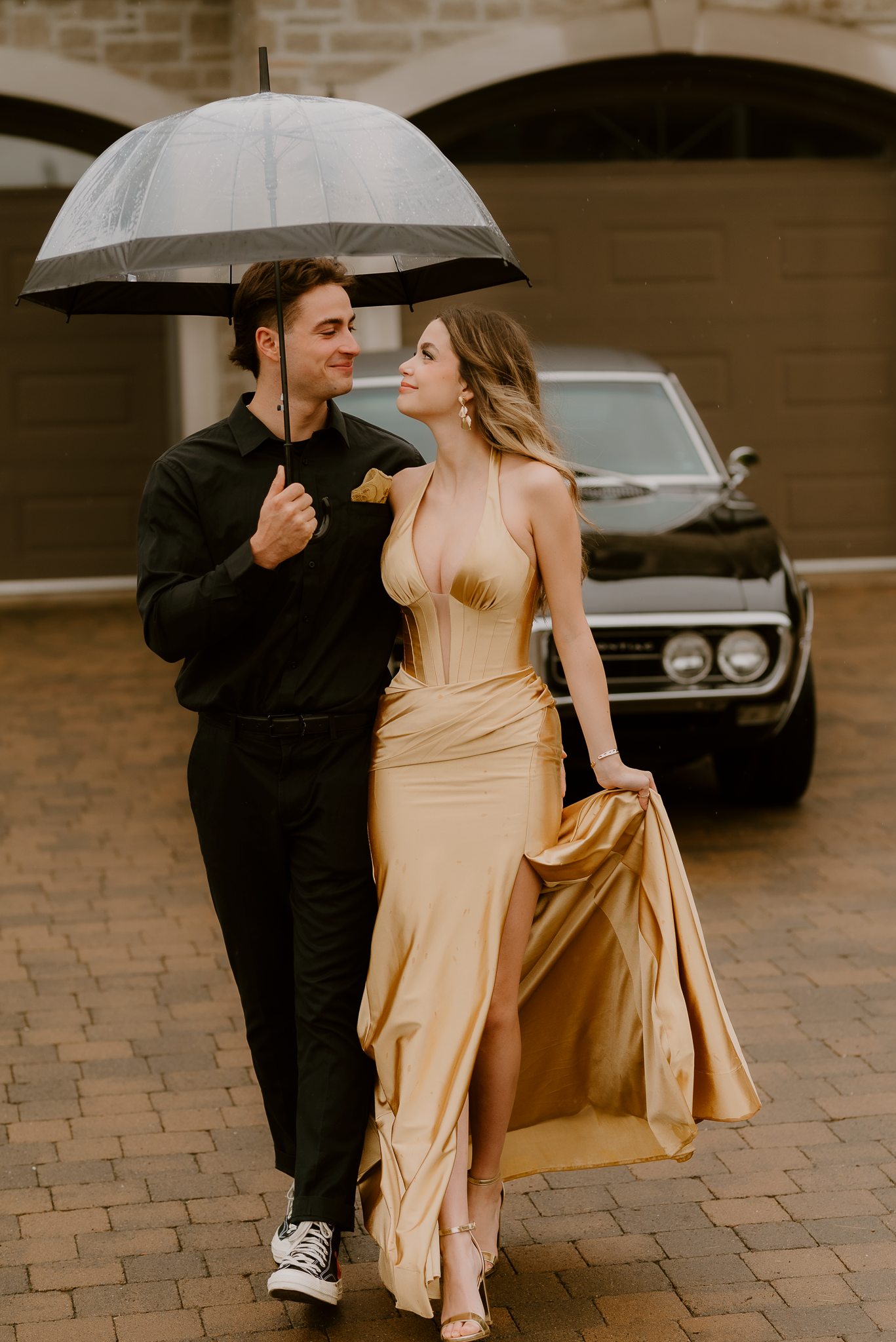 Couple walking under clear umbrella in rain, woman in gold gown, man in black, classic car background.