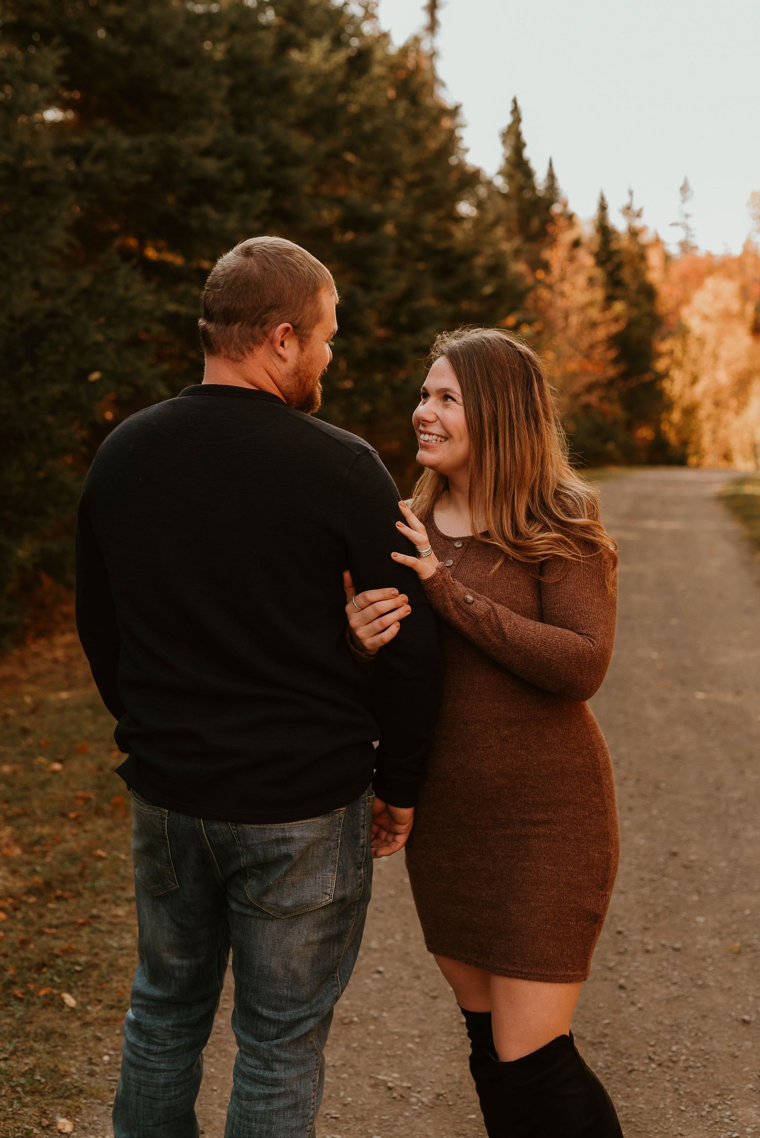 Couple smiling at each other on a tree-lined path in autumn, woman in brown sweater dress and knee-high boots.