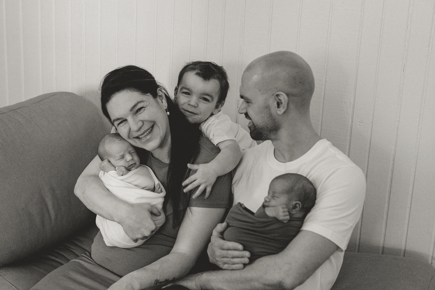 Black and white family portrait of parents holding newborn twins with toddler smiling on couch.