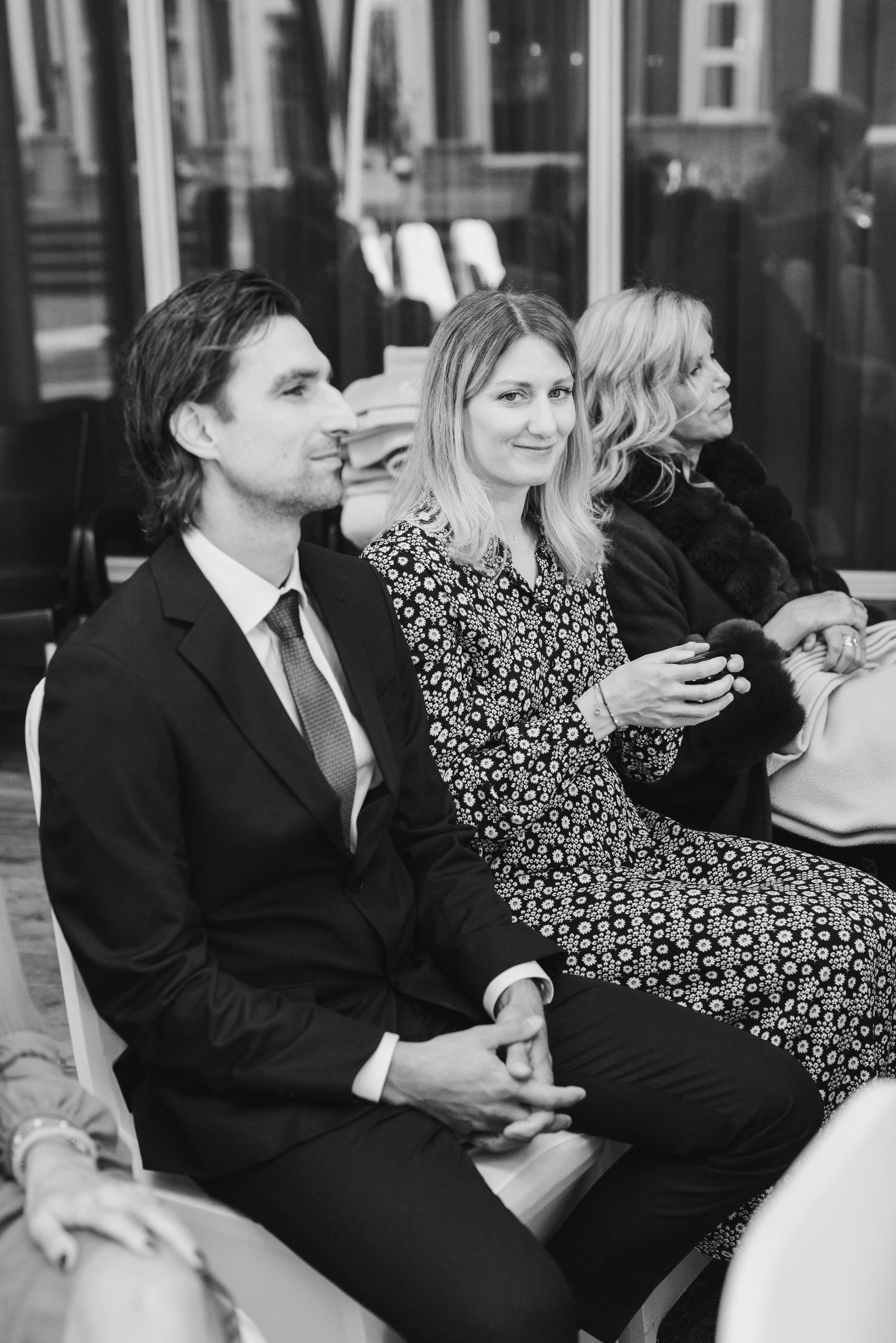 Wedding guests in formal attire seated during ceremony, with woman in patterned dress checking phone.