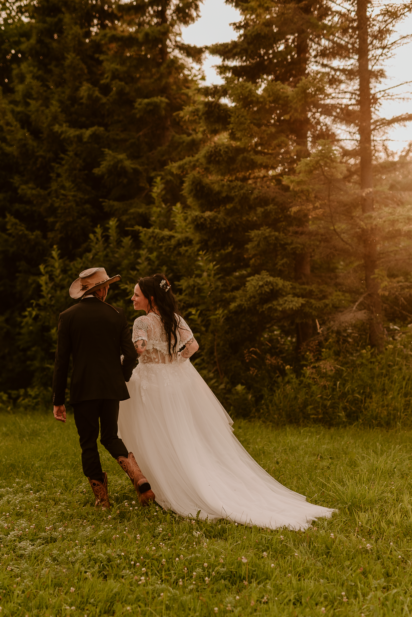 Bride and groom walking through a sunlit forest meadow, bride in flowing white gown, groom in black suit with cowboy hat.