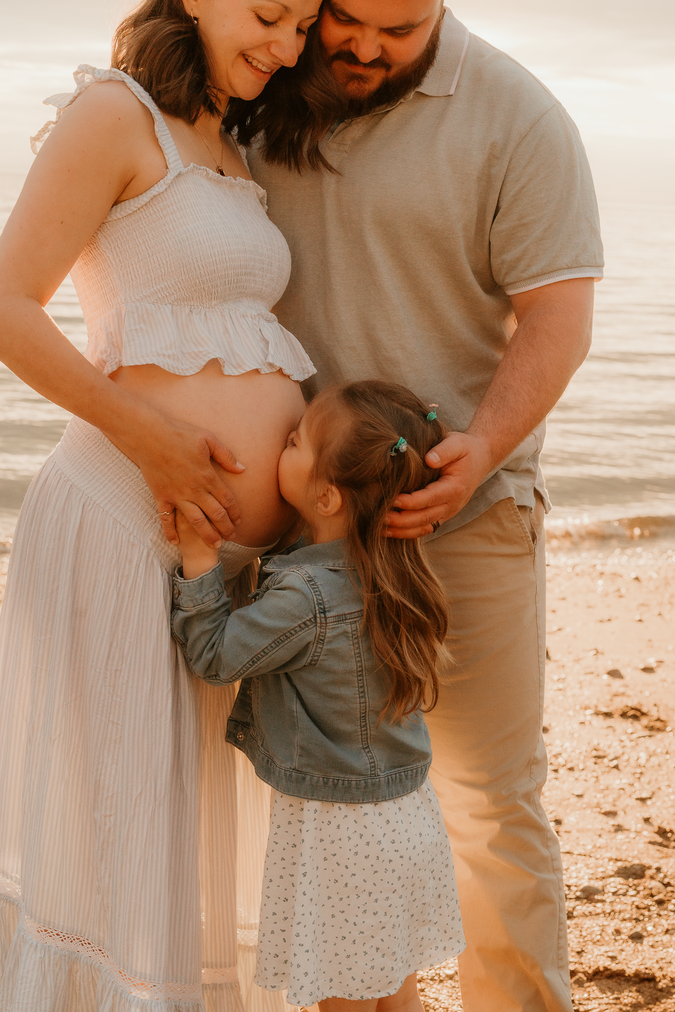 Pregnant woman in white dress with husband and young daughter kissing baby bump on sunny beach.